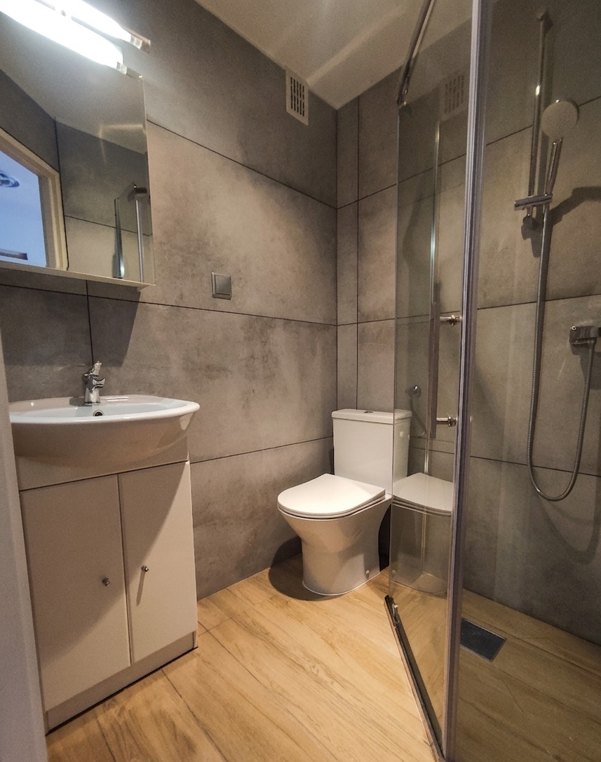 Nowoczesna łazienka z szarymi płytkami imitującymi beton, drewnopodobną podłogą, białą szafką z umywalką i kabiną prysznicową z przezroczystego szkła.