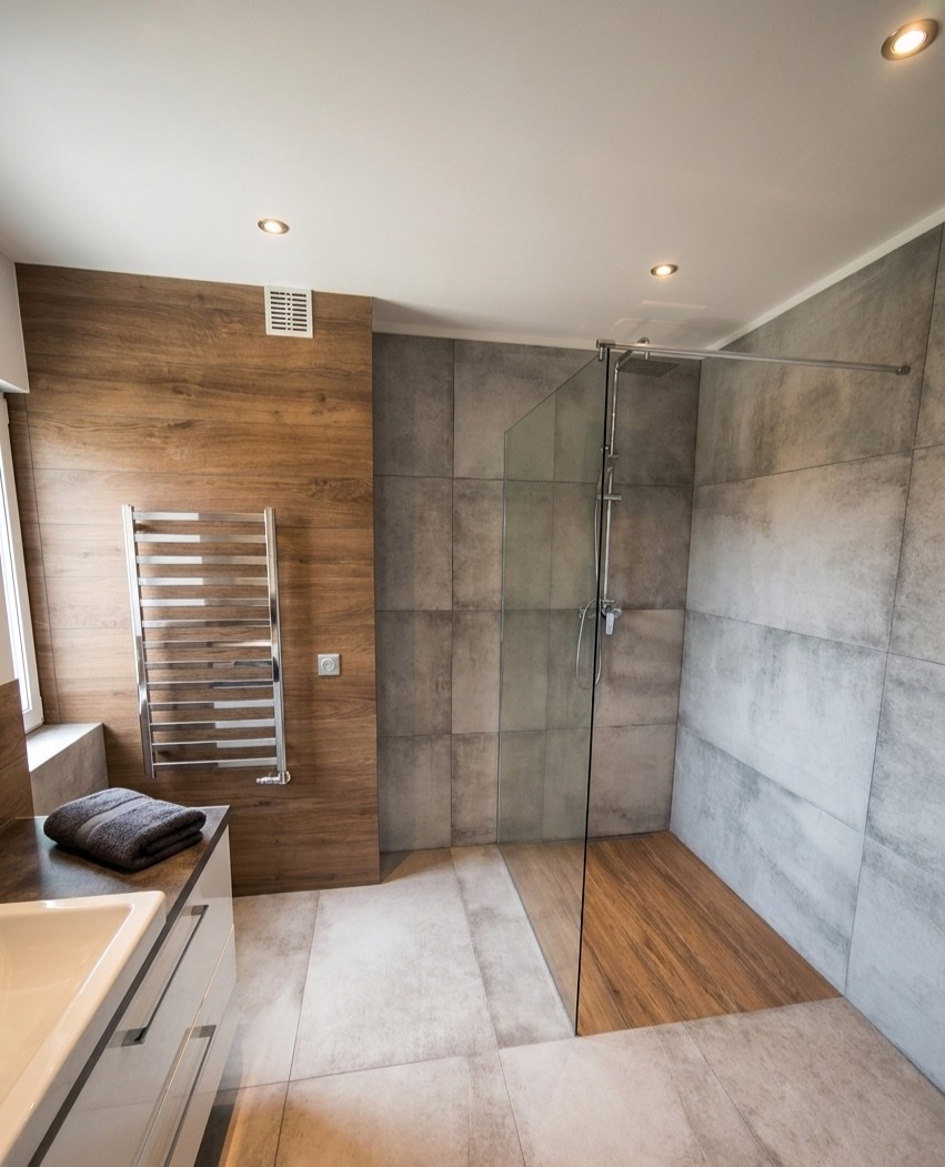 Nowoczesna łazienka z kabiną prysznicową typu walk-in, wykończona płytkami imitującymi drewno i beton. Grzejnik drabinkowy i umywalka z szafką w minimalistycznym stylu.