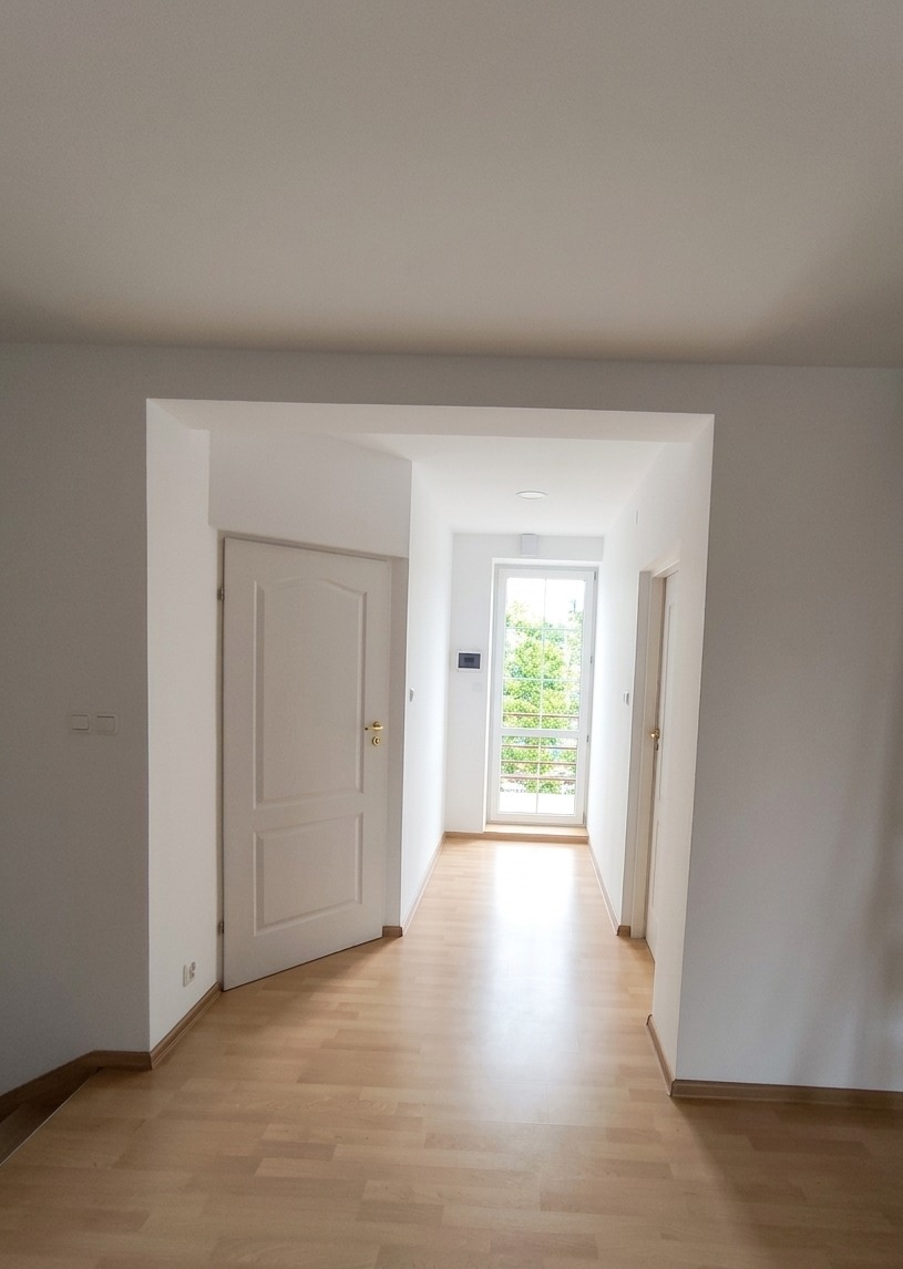 Jasne wnętrze po remoncie: białe ściany, drewniana podłoga, otwarte drzwi i okno z widokiem na zieleń. Minimalistyczny design, czystość i porządek.