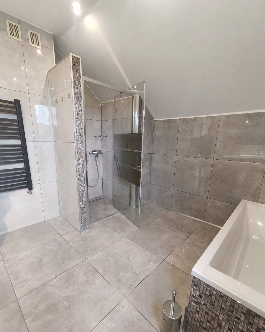 Wykończona łazienka w szarościach: kabina prysznicowa ze szklanymi drzwiami, mozaika na obrzeżach, grzejnik drabinkowy i wanna z obudową z mozaiki.