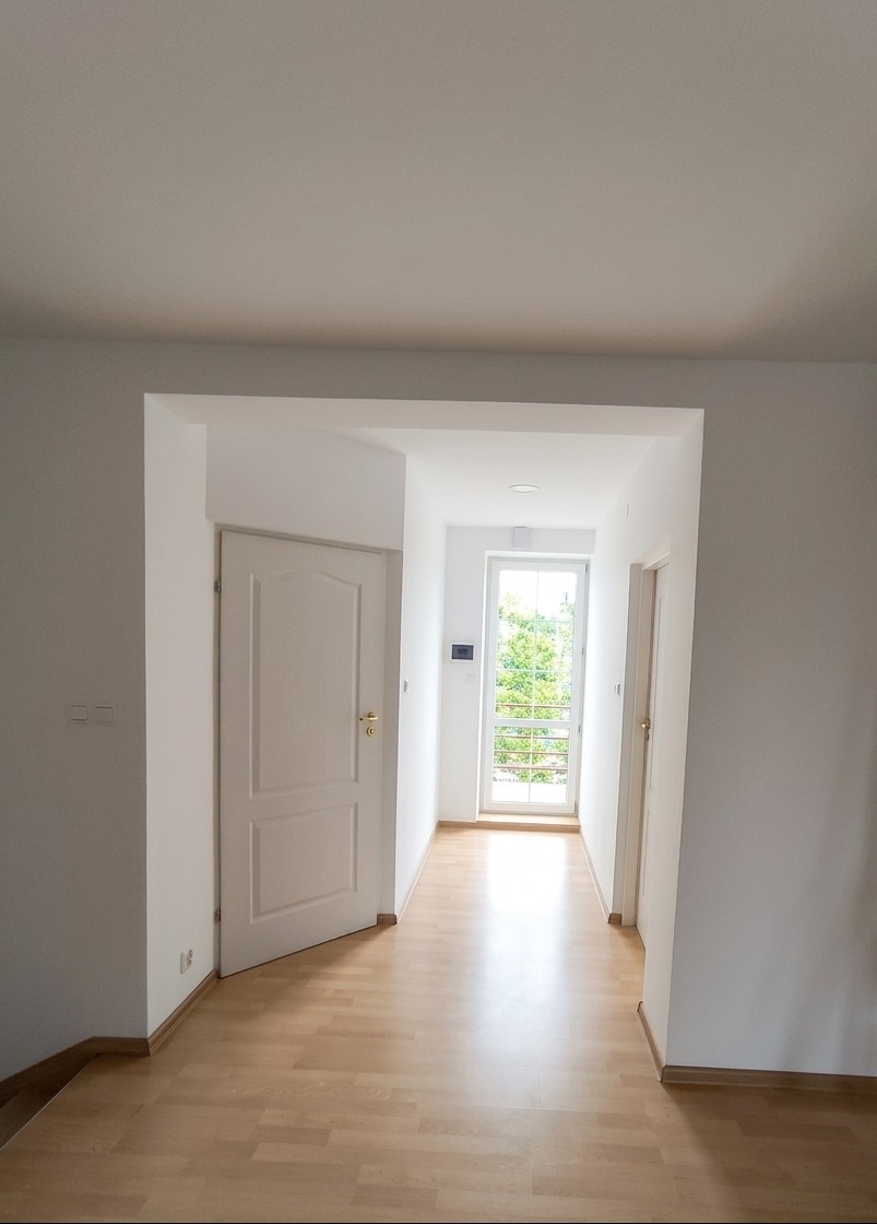 Jasne wnętrze po remoncie: białe ściany, drewniana podłoga, otwarte drzwi. Widok na korytarz z oknem i zielenią na zewnątrz. Minimalistyczny design, świeżo po wykończeniu.