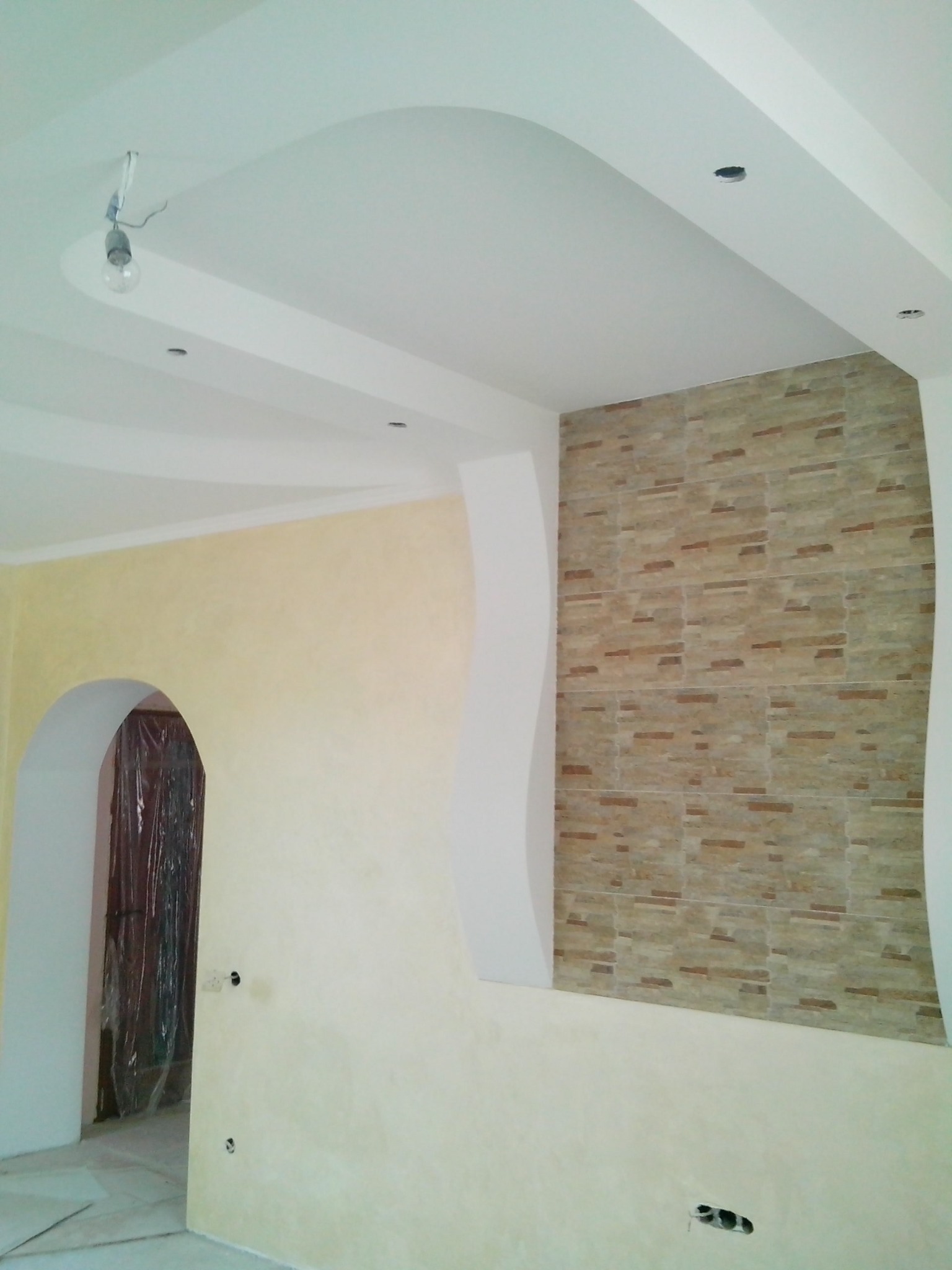 Wnętrze pokoju w trakcie remontu: pomalowane ściany w odcieniach beżu, dekoracyjna ściana z imitacji kamienia, sufit podwieszany z oświetleniem punktowym i wiszącą żarówką, drzwi zabezpieczone folią.