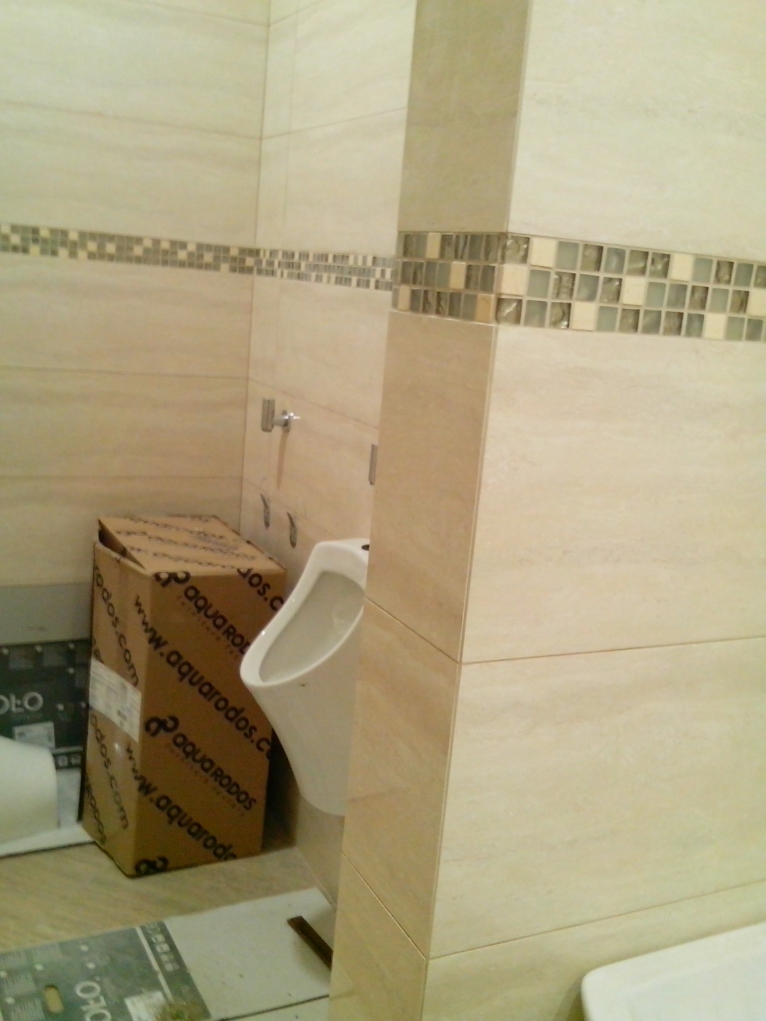Montaż pisuaru w łazience wyłożonej beżowymi płytkami z dekoracyjnym pasem mozaiki, widoczny karton z nazwą producenta armatury.