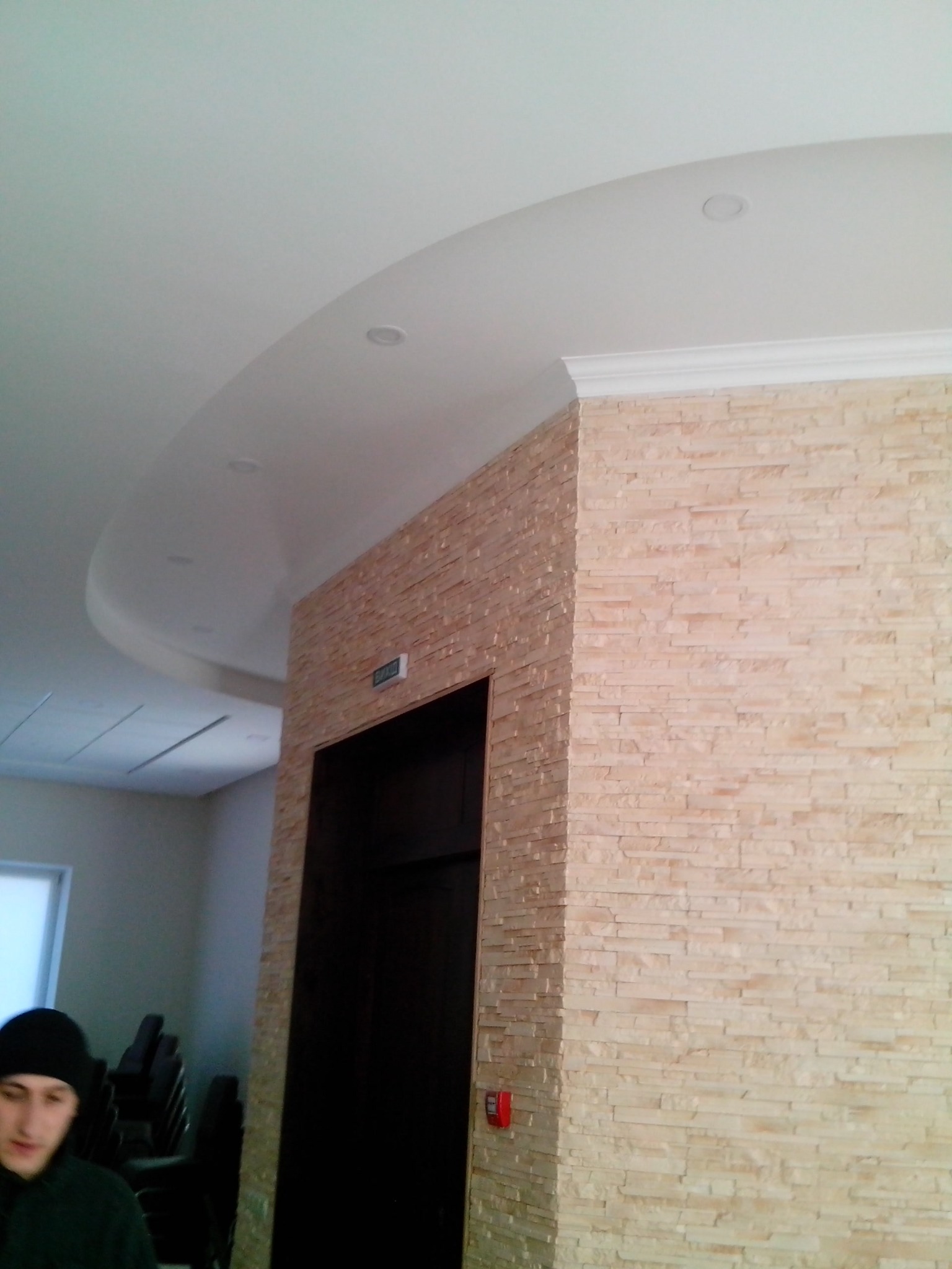 Wykończenie ściany kamieniem dekoracyjnym wewnątrz pomieszczenia, widoczny fragment sufitu podwieszanego z oświetleniem punktowym oraz ciemne drzwi.