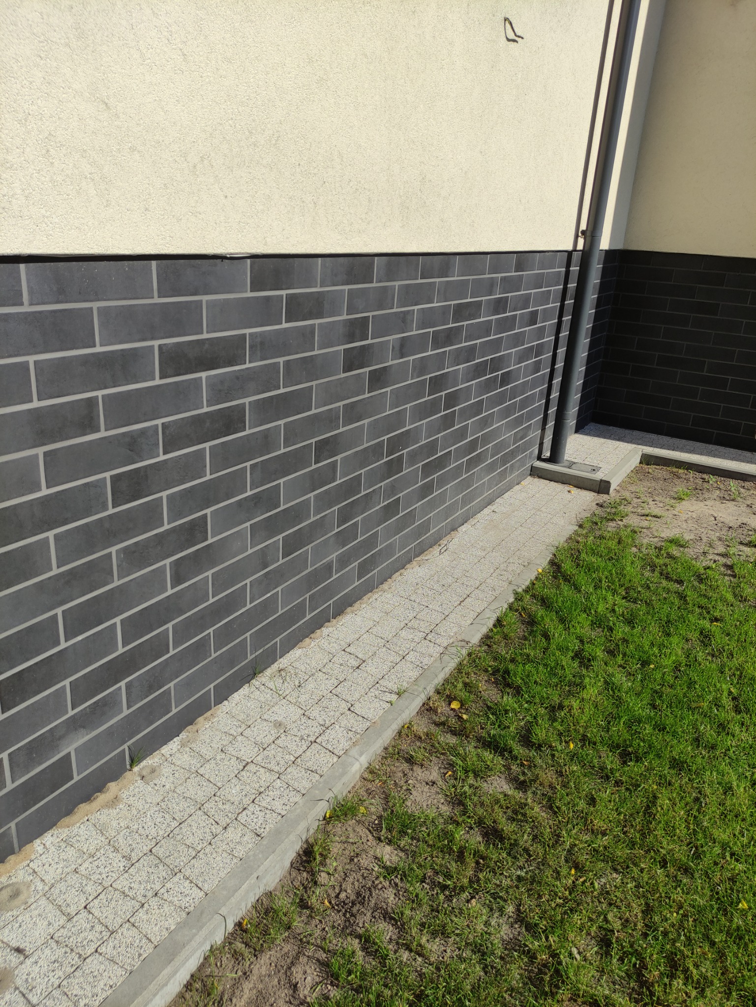 Elewacja budynku z ciemnoszarą okładziną imitującą cegłę, jasny tynk powyżej, chodnik z kostki brukowej i fragment trawnika.