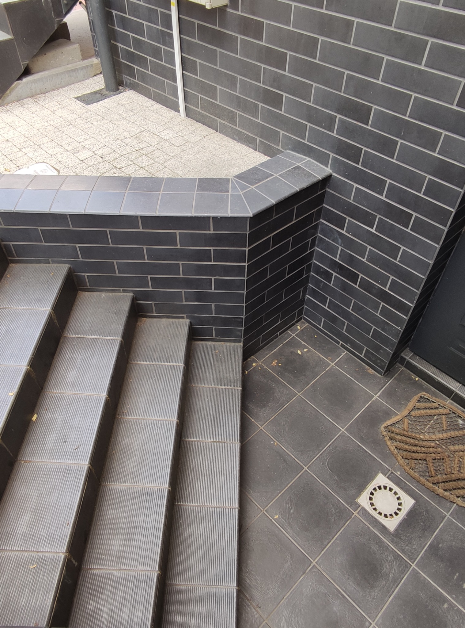 Zewnętrzne schody wyłożone ciemnoszarą płytką z ryflowaną powierzchnią, obok ściana z ciemnej cegły z jasnymi spoinami i fragment chodnika z jasnej kostki.