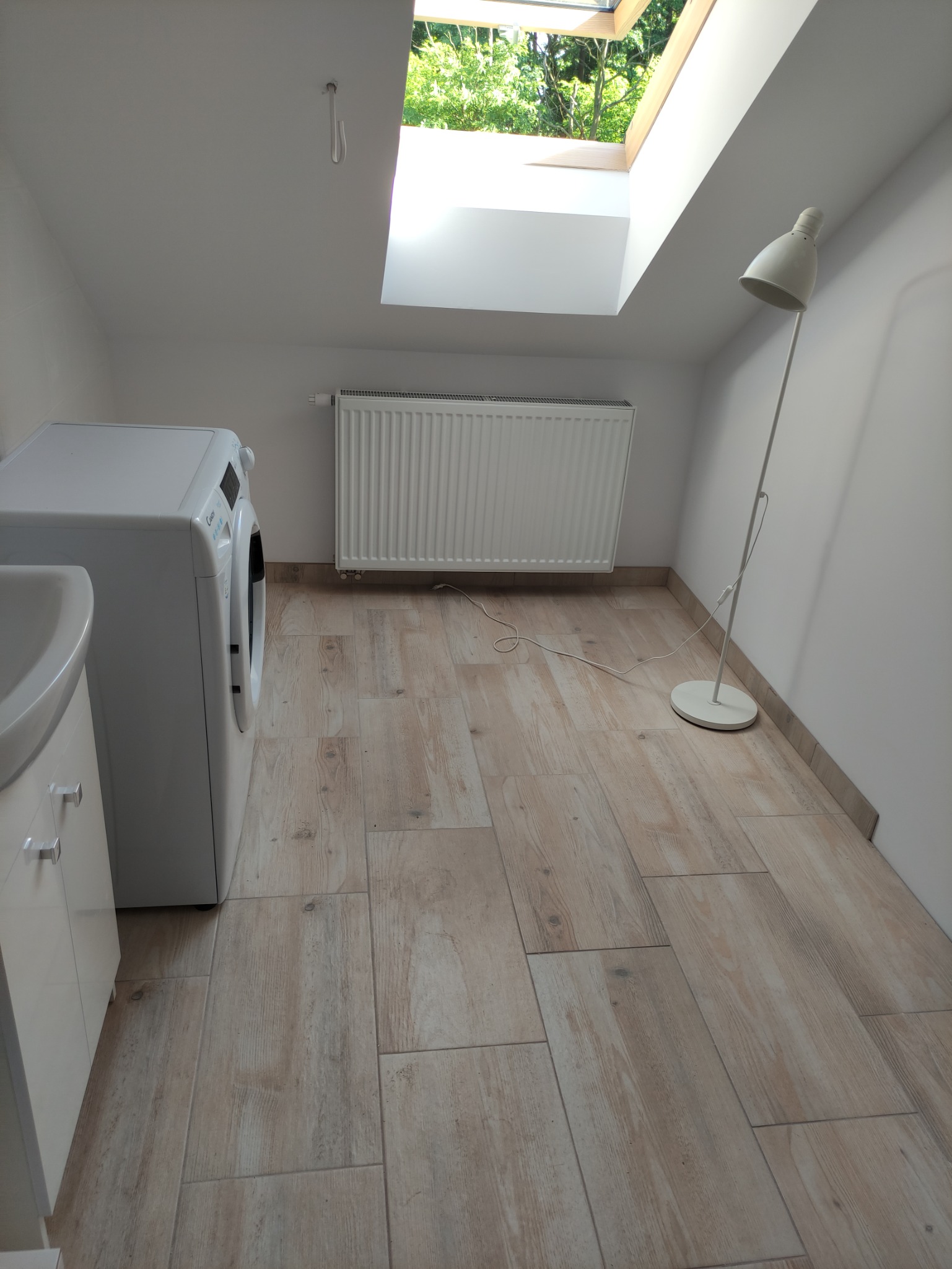 Jasna łazienka na poddaszu z pralką, umywalką, grzejnikiem, lampą stojącą i oknem dachowym z widokiem na zieleń.