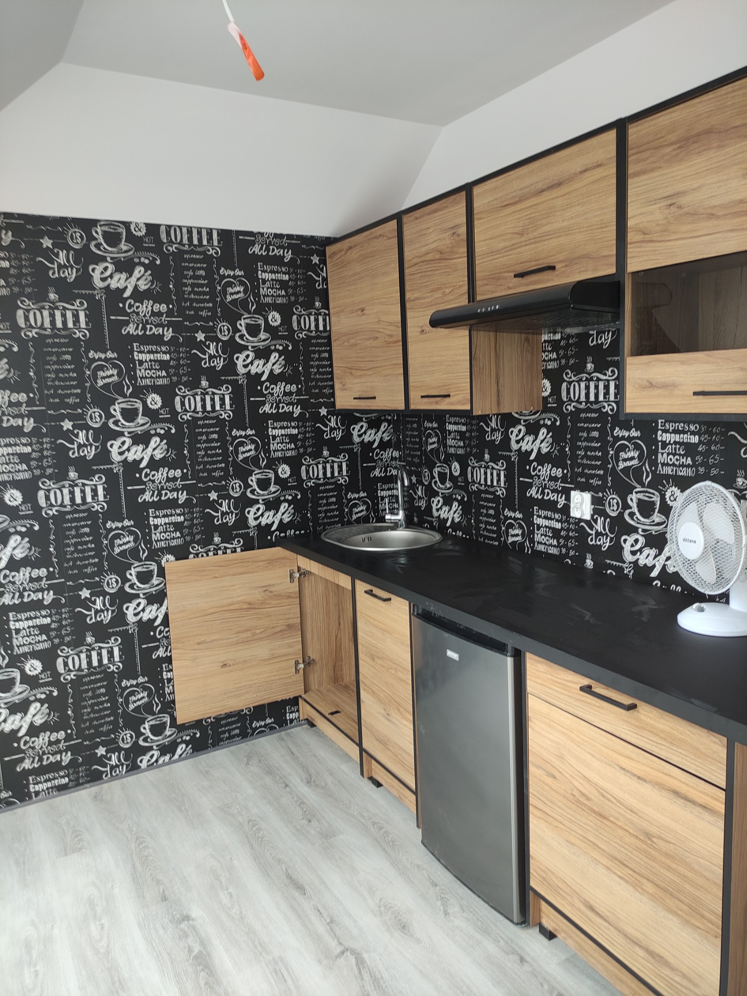 Wykończona kuchnia z drewnianymi szafkami, czarnym blatem, tapetą z motywem kawy i małą srebrną lodówką pod blatem. W rogu stoi biały wentylator.