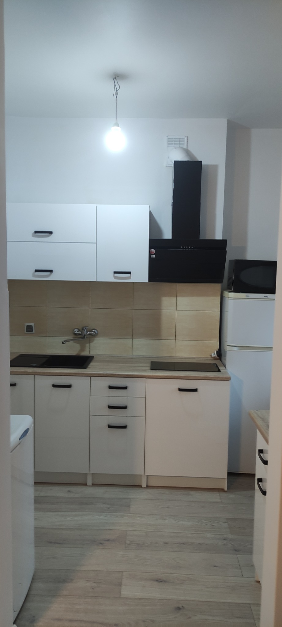 Nowoczesna kuchnia z białymi szafkami, czarnym okapem i drewnianą podłogą, widziana z perspektywy wejścia.