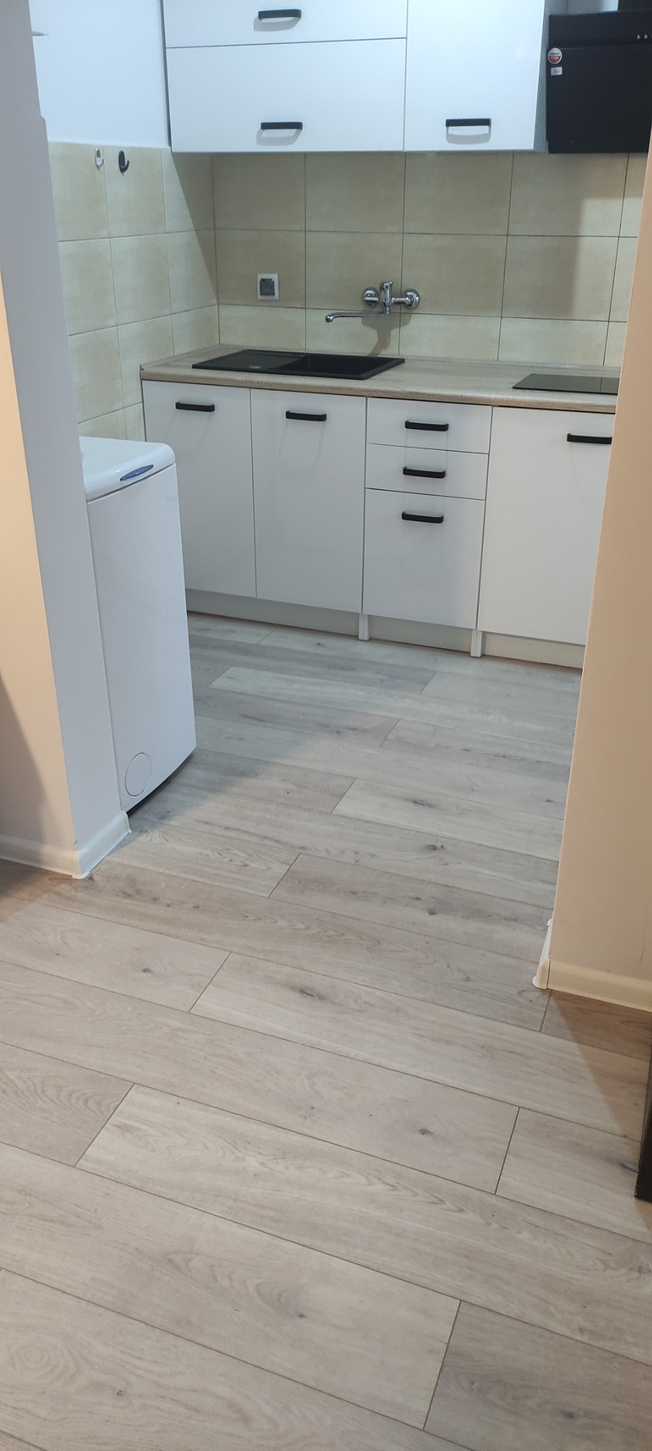 Nowoczesna kuchnia z białymi szafkami z czarnymi uchwytami, jasnymi płytkami na ścianie i panelami podłogowymi, pralka w rogu