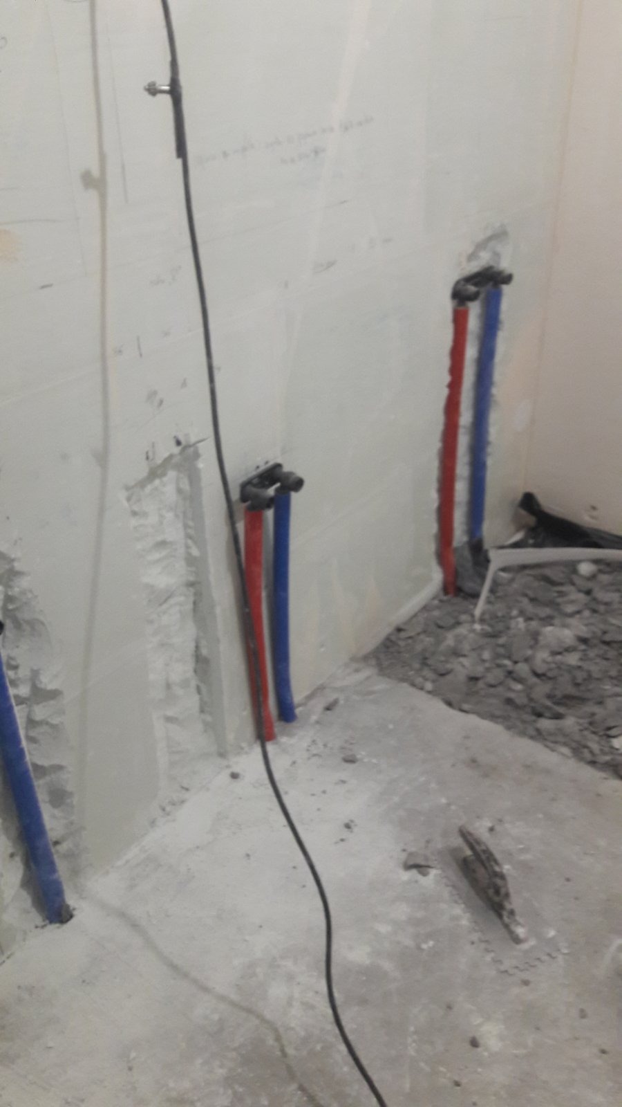 Przygotowanie instalacji hydraulicznej w łazience: widoczne rury doprowadzające wodę (czerwona i niebieska) wystające ze ściany, fragment ściany z uszkodzonym tynkiem i gruz na podłodze.