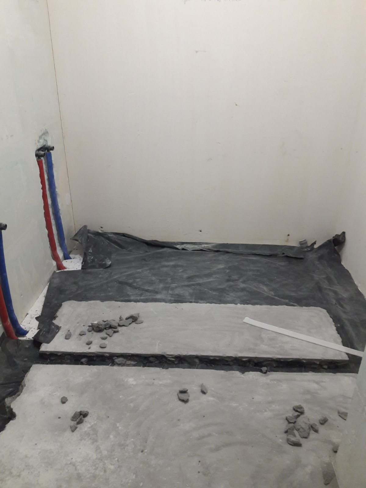 Przygotowanie podłoża w łazience: widoczne rury wodne w ścianie, rozłożona czarna folia ochronna i betonowa wylewka z gruzem.