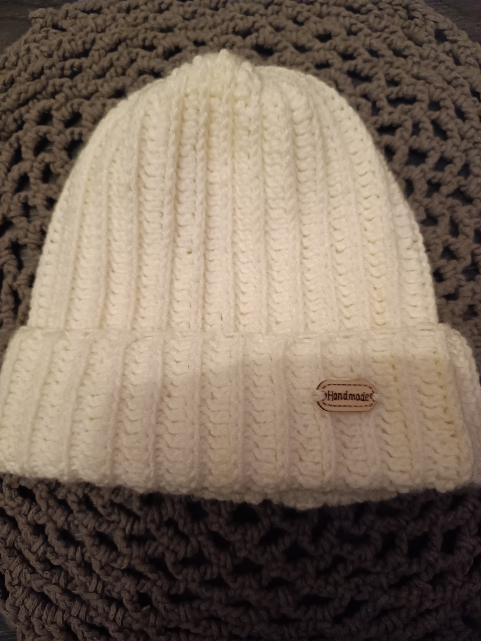 Kremowa czapka z grubymi prążkami, z naszywką 'Handmade', leżąca na szarym, dzierganym tle.