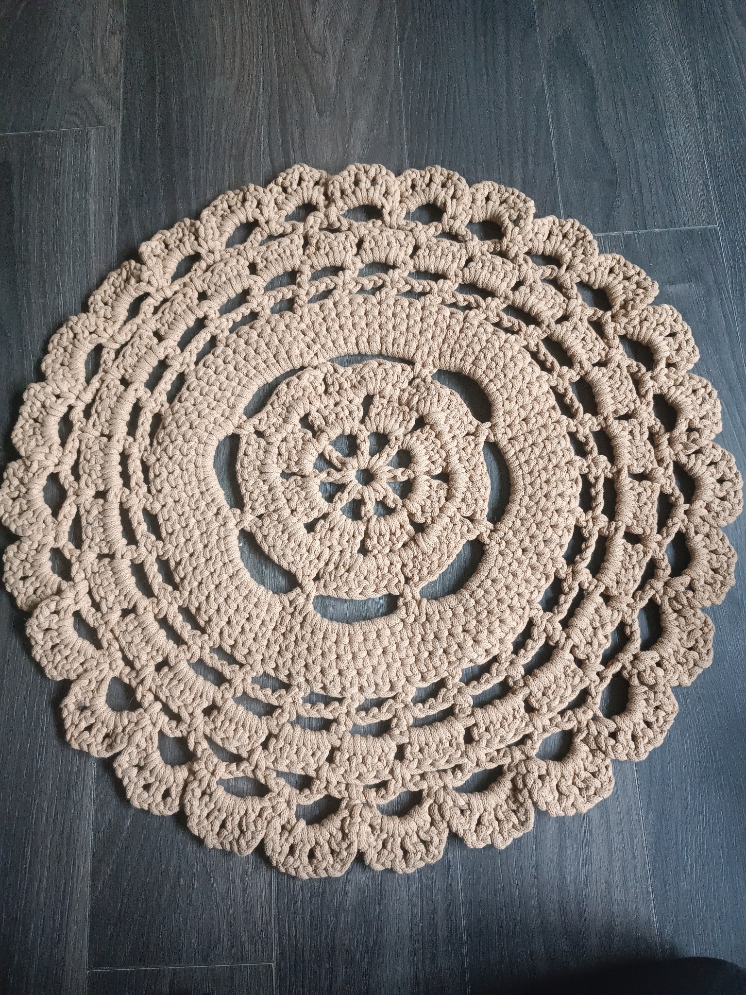 Beżowa, okrągła serwetka z grubego sznurka, wykonana na szydełku, leży na ciemnoszarej podłodze z paneli.