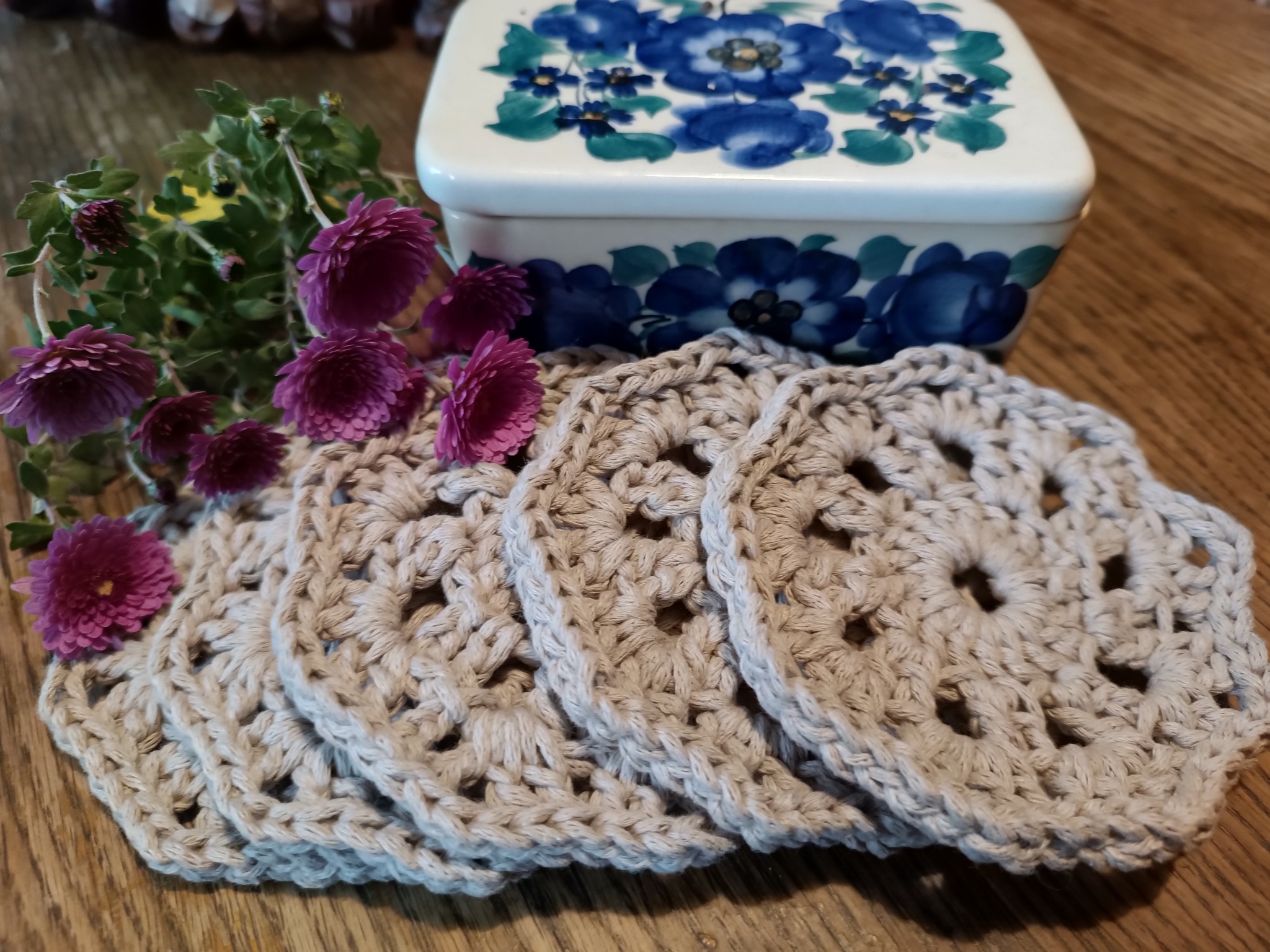 Stos ręcznie robionych, beżowych, ażurowych podkładek pod kubki z włóczki, ozdobionych fioletowymi chryzantemami, obok ceramicznego pudełka z motywem niebieskich kwiatów, na drewnianym stole.
