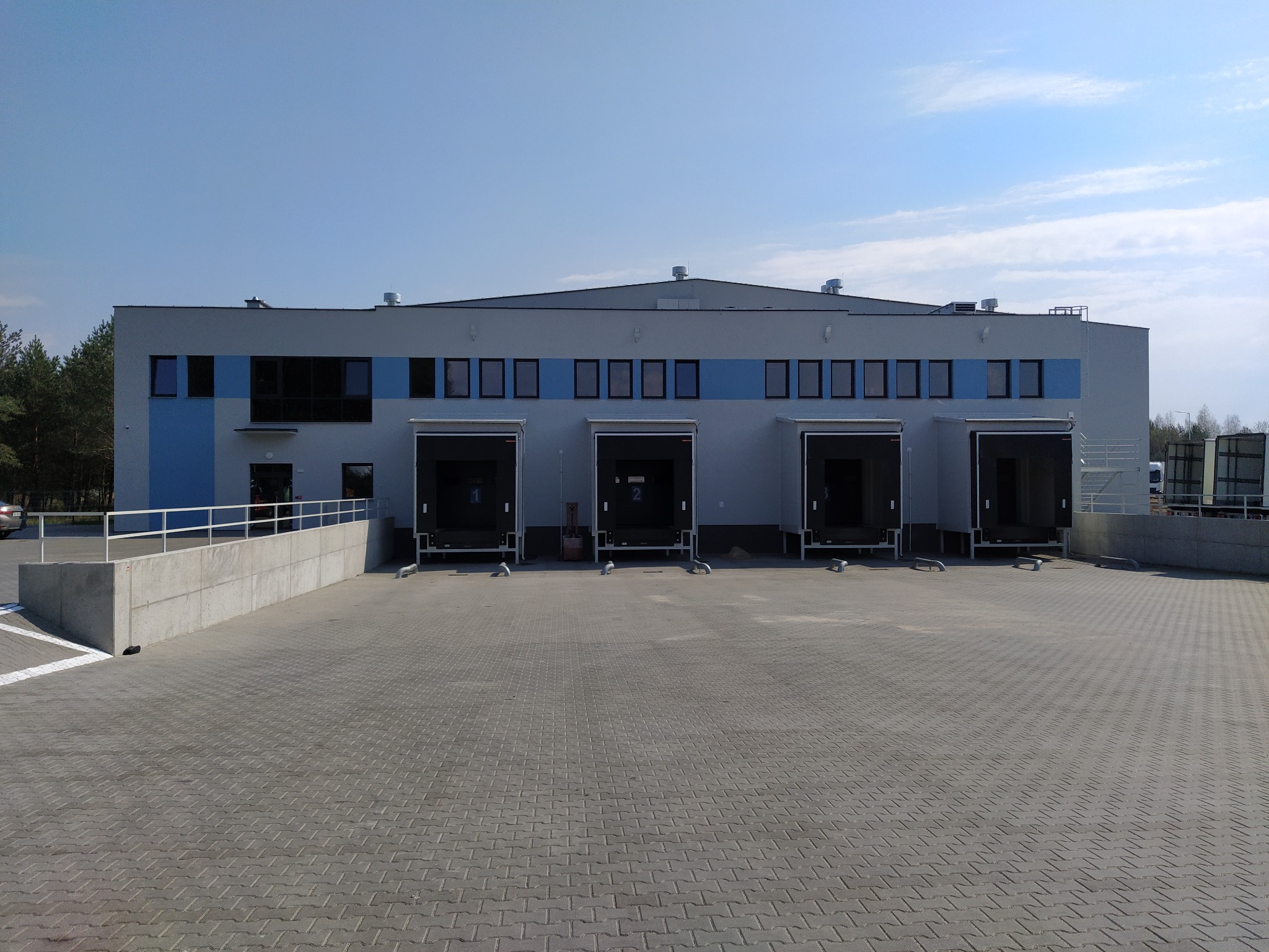 Nowoczesny budynek magazynowy z czterema bramami załadunkowymi i betonową rampą, widok z zewnątrz w słoneczny dzień.