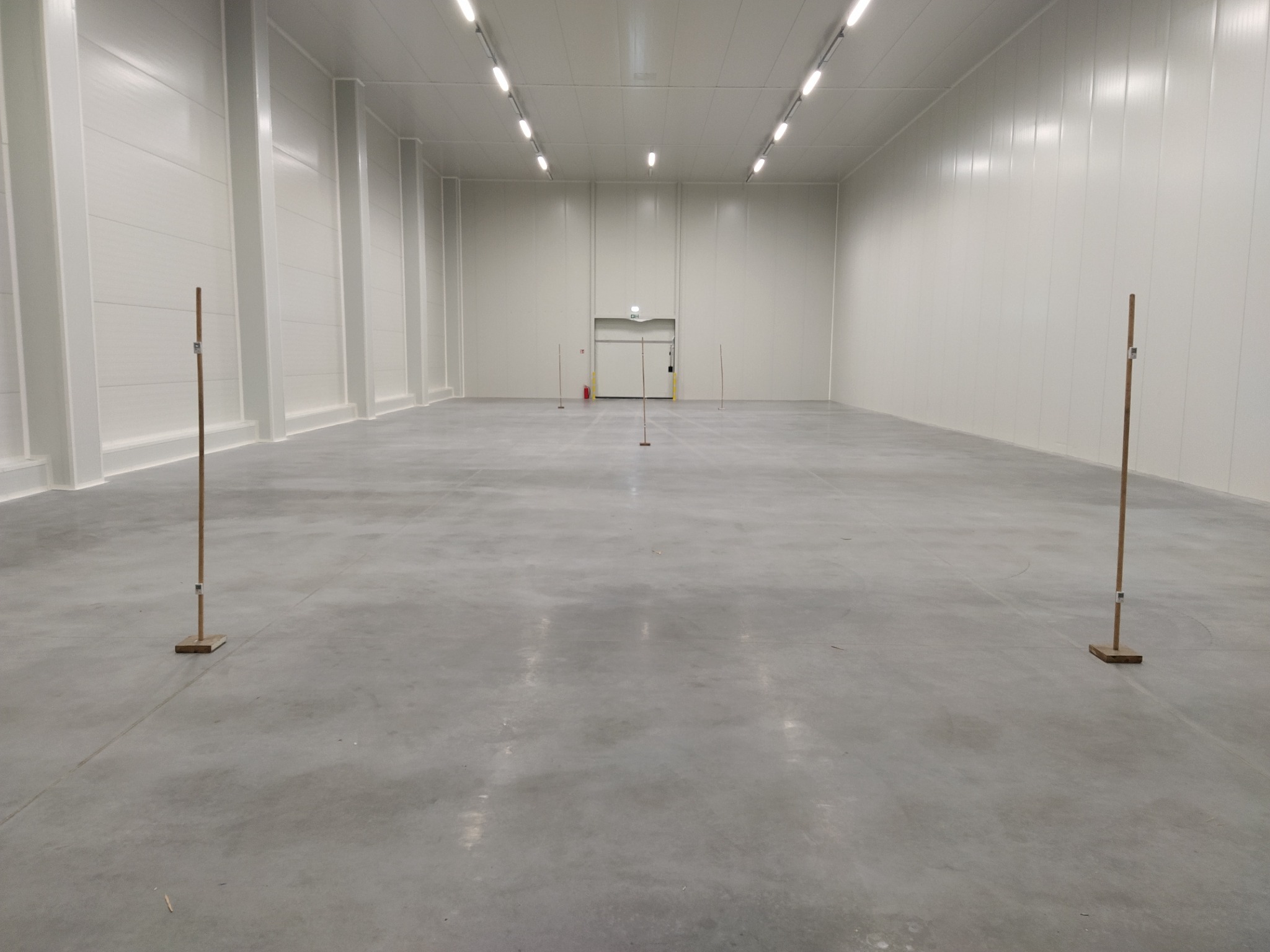 Pusta hala magazynowa z betonową podłogą, białymi ścianami z paneli i rzędem lamp sufitowych; widoczne drewniane znaczniki na podłodze.