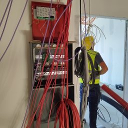 IEM Oleksandr Oleksiienko - Rozdzielnia elektryczna z licznymi przewodami w kolorach czerwonym i fioletowym, instalowana przez pracownika w żółtym kasku i kamizelce odblaskowej w surowym wnętrzu.