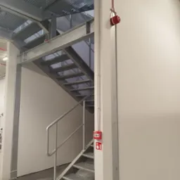 Metalowe schody ewakuacyjne z kratownicowymi stopniami i barierką, widoczny sygnalizator pożarowy na ścianie i przewody elektryczne w listwach.
