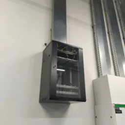 Zamontowana na białej ścianie szafa rack z oszklonymi drzwiami, z widocznymi kablami wewnątrz. Nad szafą pionowy kanał kablowy, a obok pionowe koryta kablowe. Widoczny również fragment białej szafy...