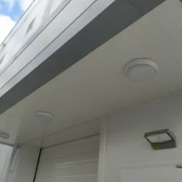 Widok z dołu na fragment elewacji budynku z trzema okrągłymi lampami sufitowymi i jedną prostokątną lampą ścienną, białe panele elewacyjne z szarymi akcentami, błękitne niebo w tle.