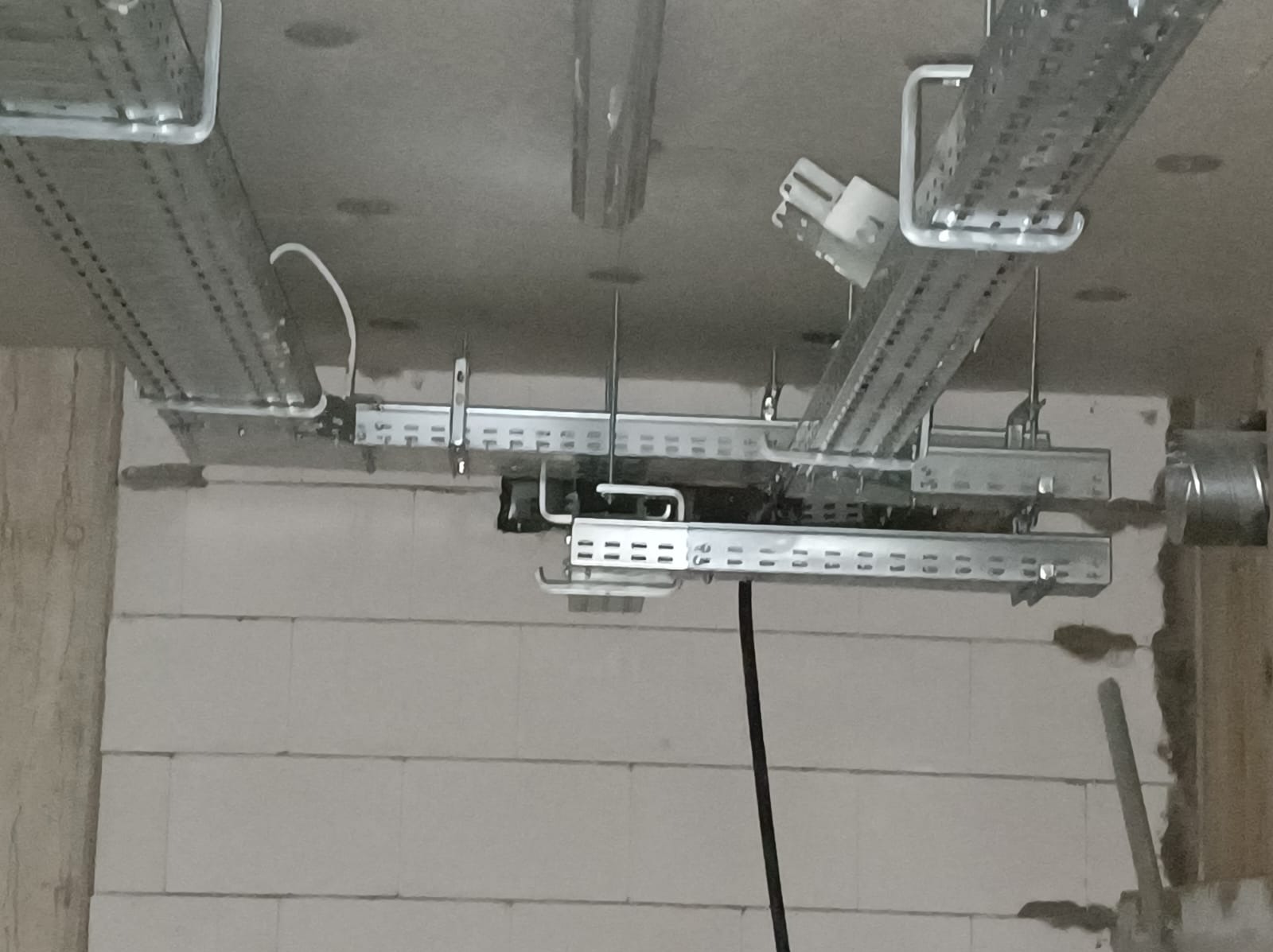 Metalowe korytka kablowe zawieszone pod sufitem w surowym stanie, widoczne przewody i mocowania, ściany z białych bloczków.