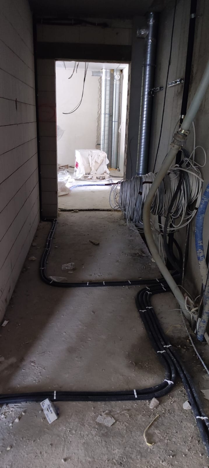Kable elektryczne w peszlach ułożone na podłodze w surowym stanie, widoczne rury wentylacyjne i materiały budowlane w tle, ściany w trakcie wykończenia.