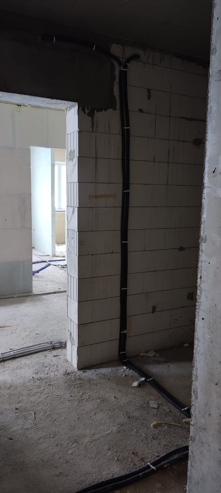 Surowy stan wnętrza z widocznymi przewodami elektrycznymi prowadzonymi po ścianie z bloczków betonowych, zabezpieczonymi białymi uchwytami, oraz rurami instalacyjnymi na podłodze.