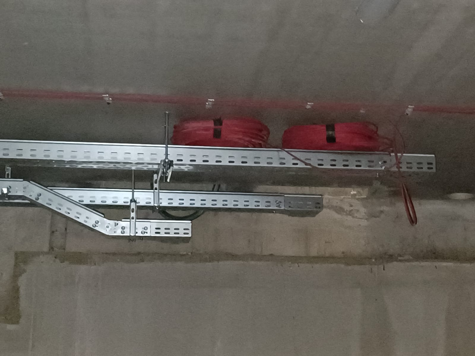 Metalowe korytka kablowe z perforacją, zawieszone pod betonowym sufitem, zawierające zwitki czerwonych przewodów elektrycznych.