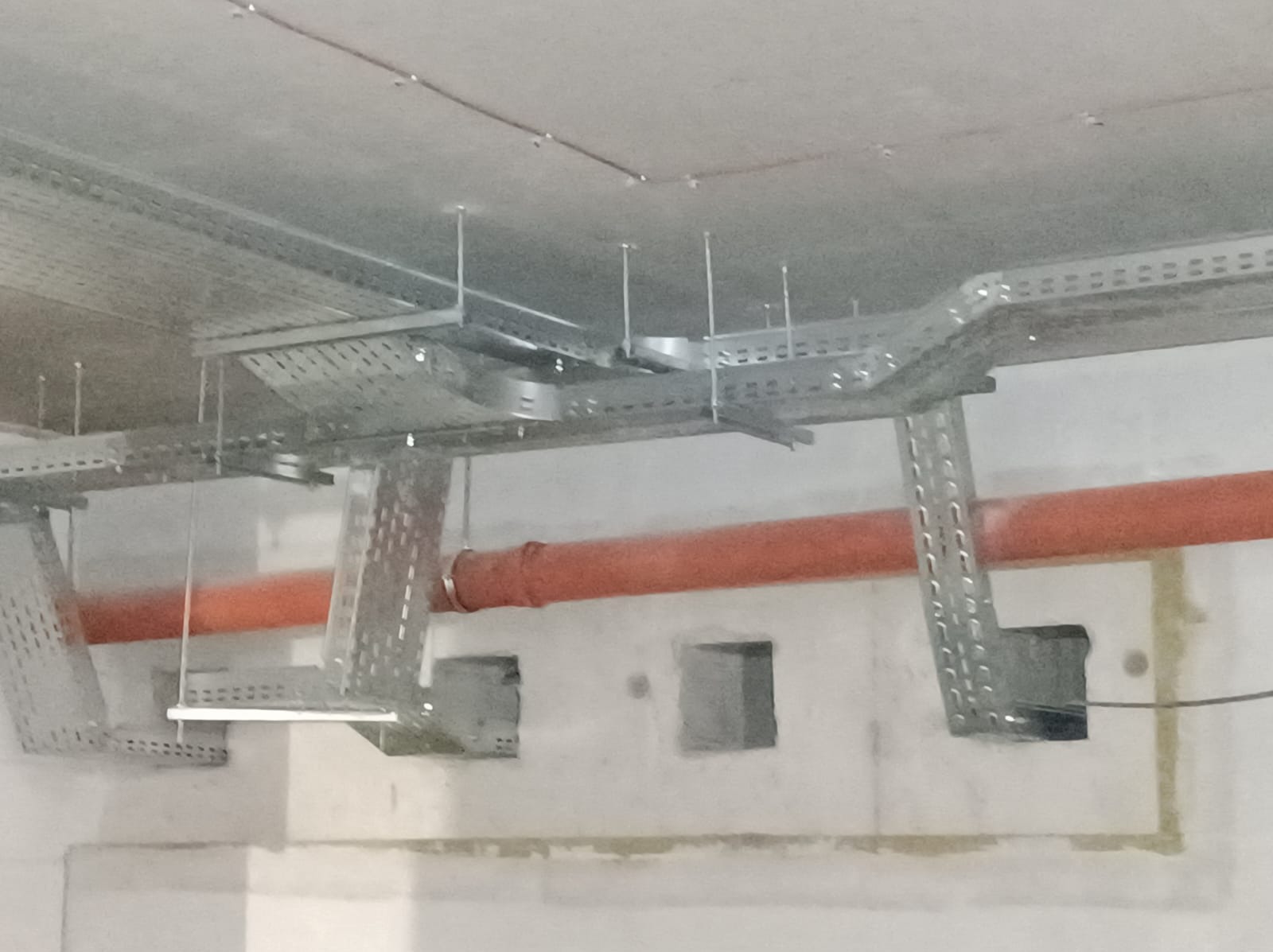 Metalowe korytka kablowe zawieszone pod sufitem, widoczne otwory w ścianie i pomarańczowa rura w tle.