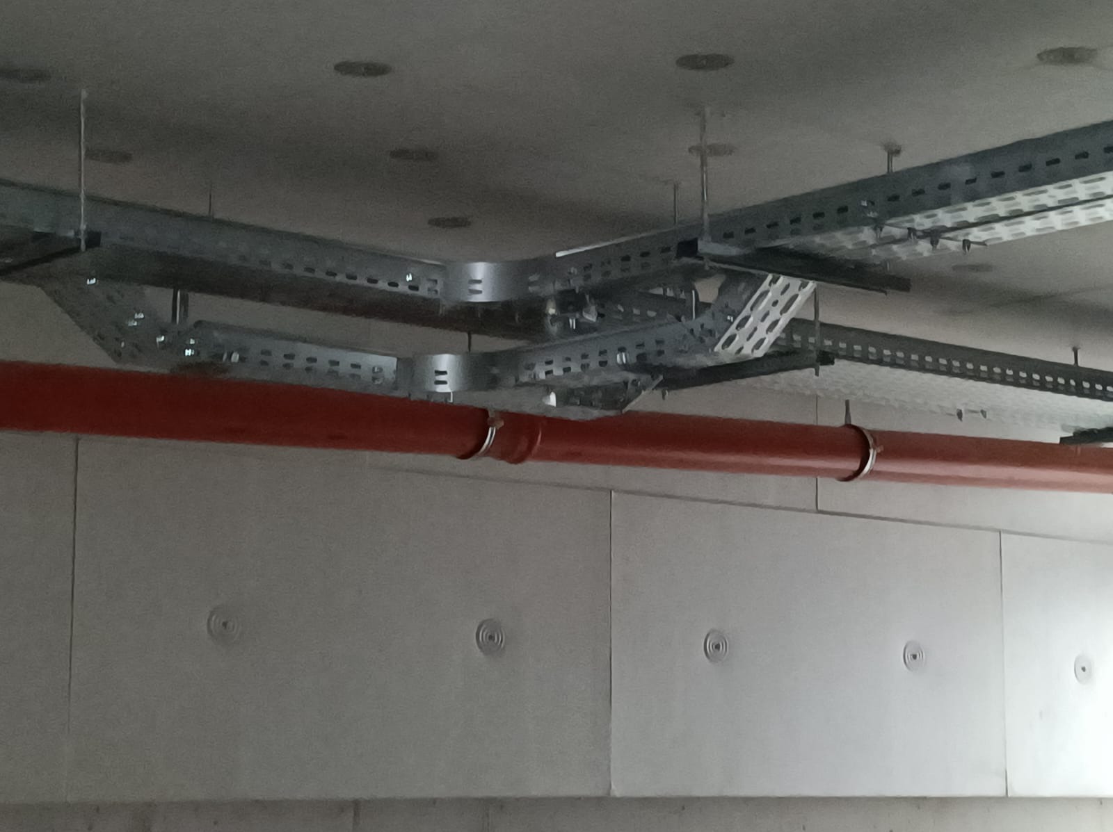 Szare koryta kablowe pod sufitem, z widocznym pomarańczowym rurociągiem mocowanym do ściany z betonu architektonicznego.