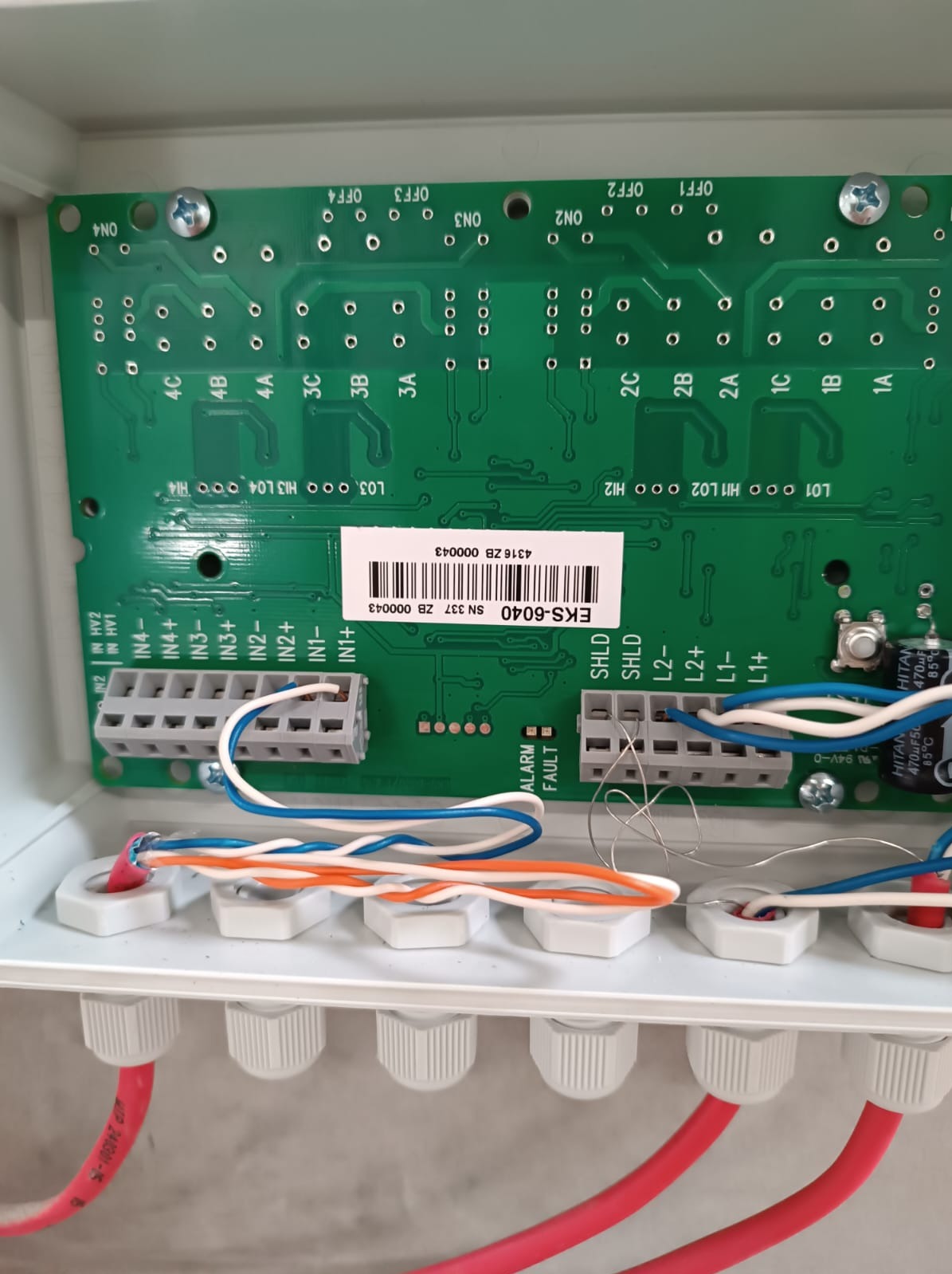 Wnętrze skrzynki z zieloną płytką drukowaną, liczne złącza, podłączone kable w kolorach białym, pomarańczowym, niebieskim i czerwonym, widoczna etykieta z kodem kreskowym EKS-6040.