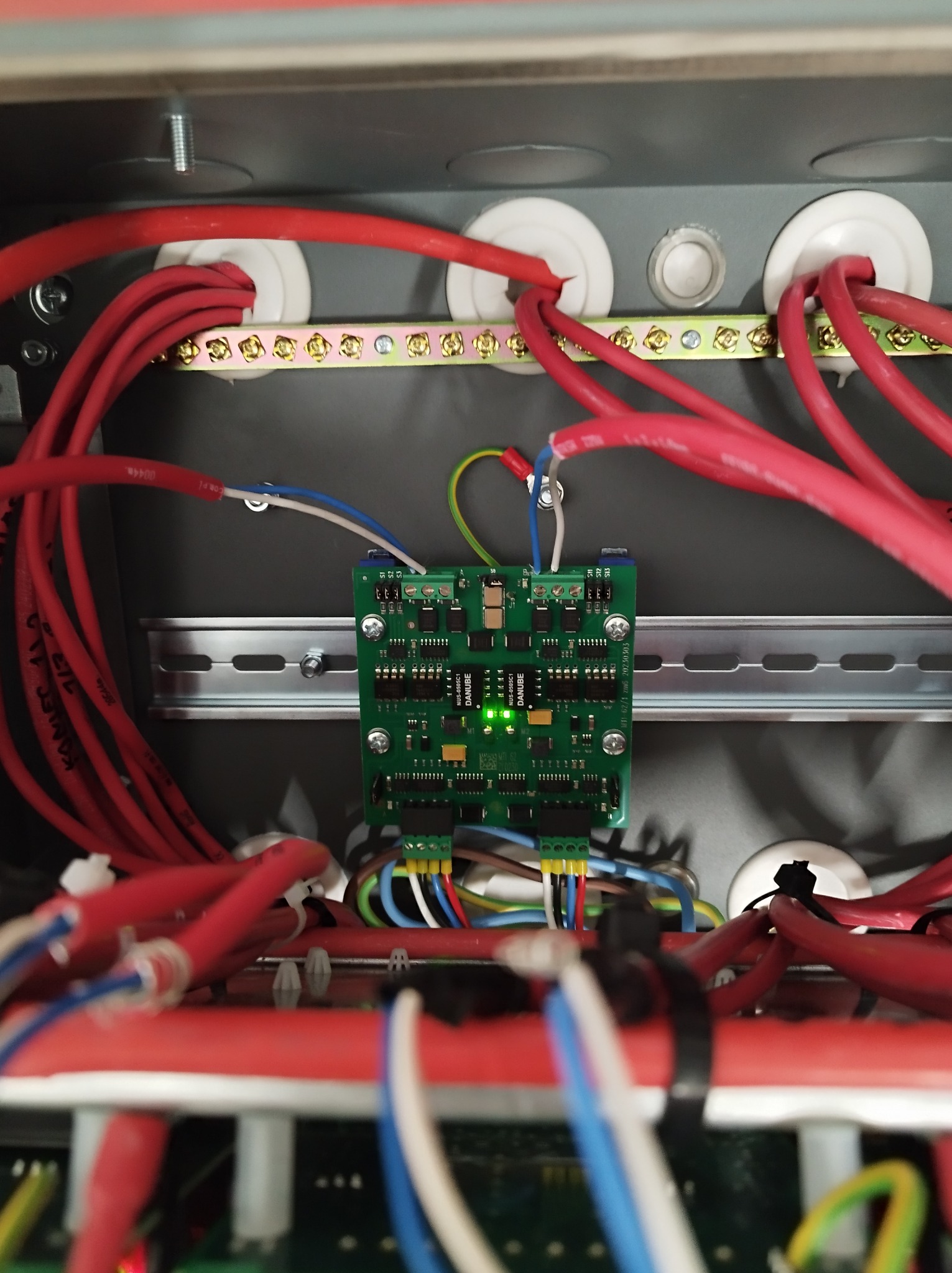 Wnętrze szarej skrzynki rozdzielczej z czerwonymi przewodami, listwą zaciskową, zieloną płytką drukowaną Danube z diodą LED oraz różnokolorowymi przewodami podłączonymi do złączek.