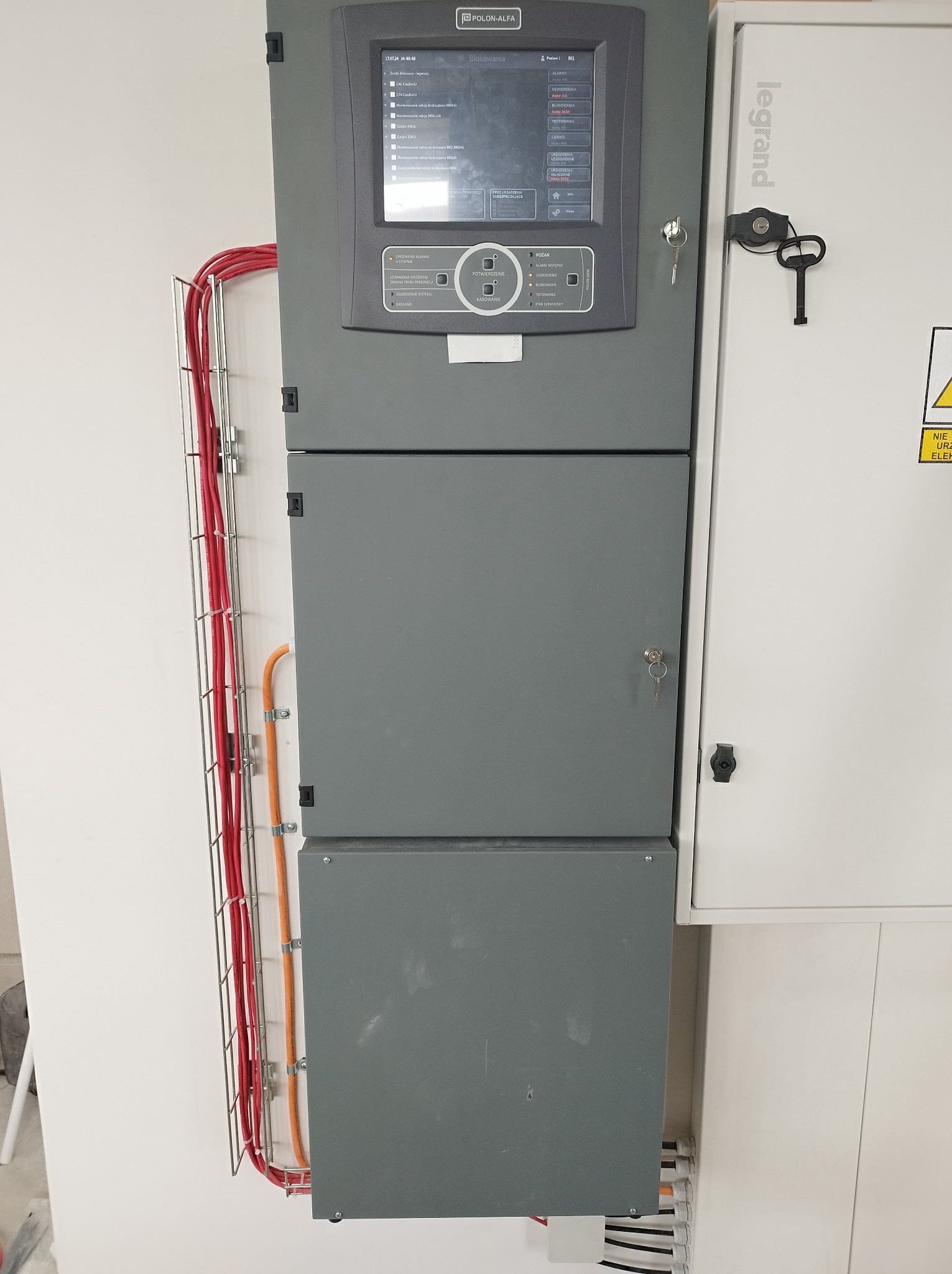 Szara szafa sterownicza Polon-Alfa z wyświetlaczem i kluczykiem, obok biała szafa Legrand z kluczem, kable w korytkach na ścianie.