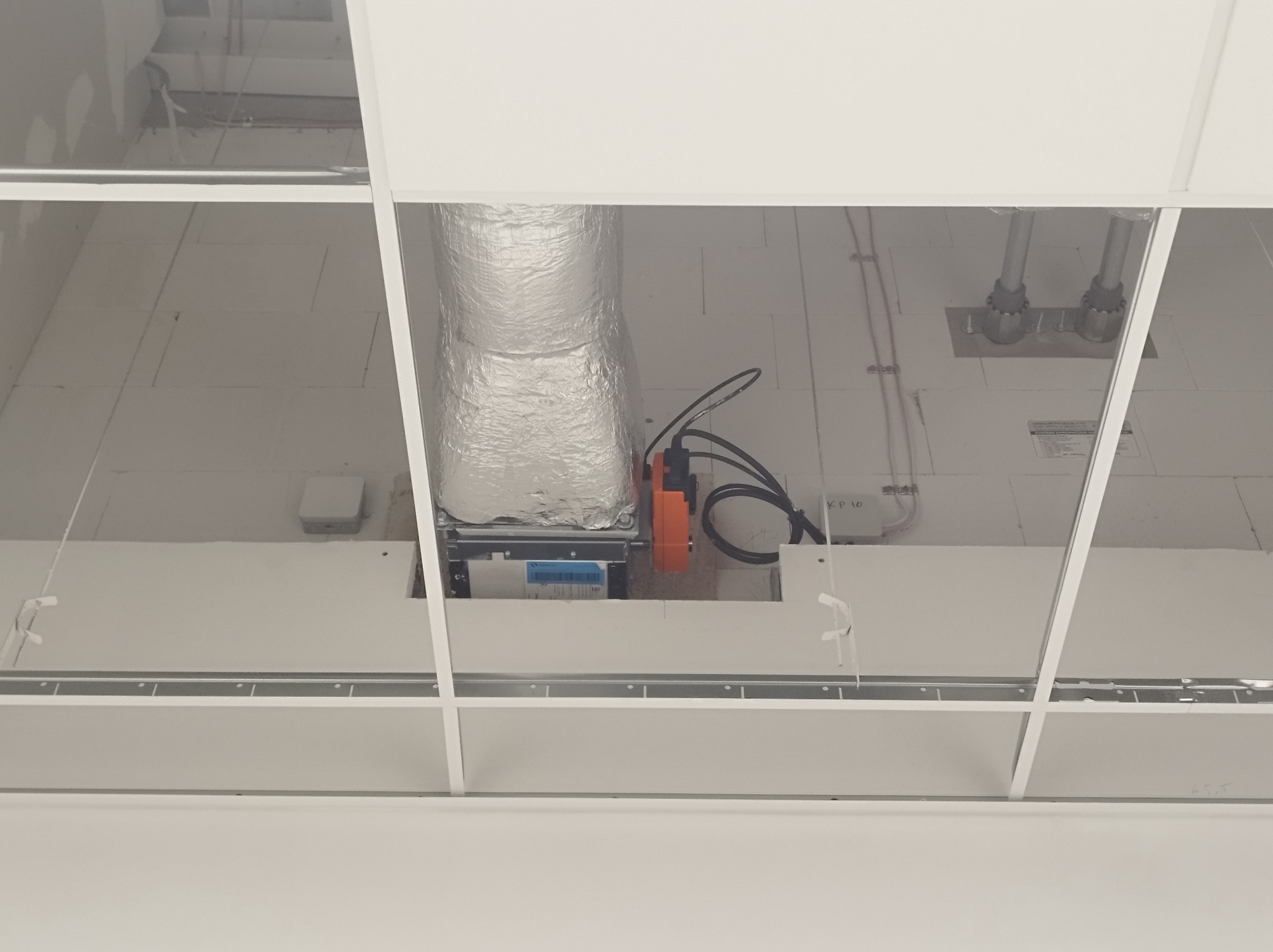 Widok z dołu na sufit podwieszany z widoczną instalacją wentylacyjną, rurami i przewodami elektrycznymi w przestrzeni międzystropowej.