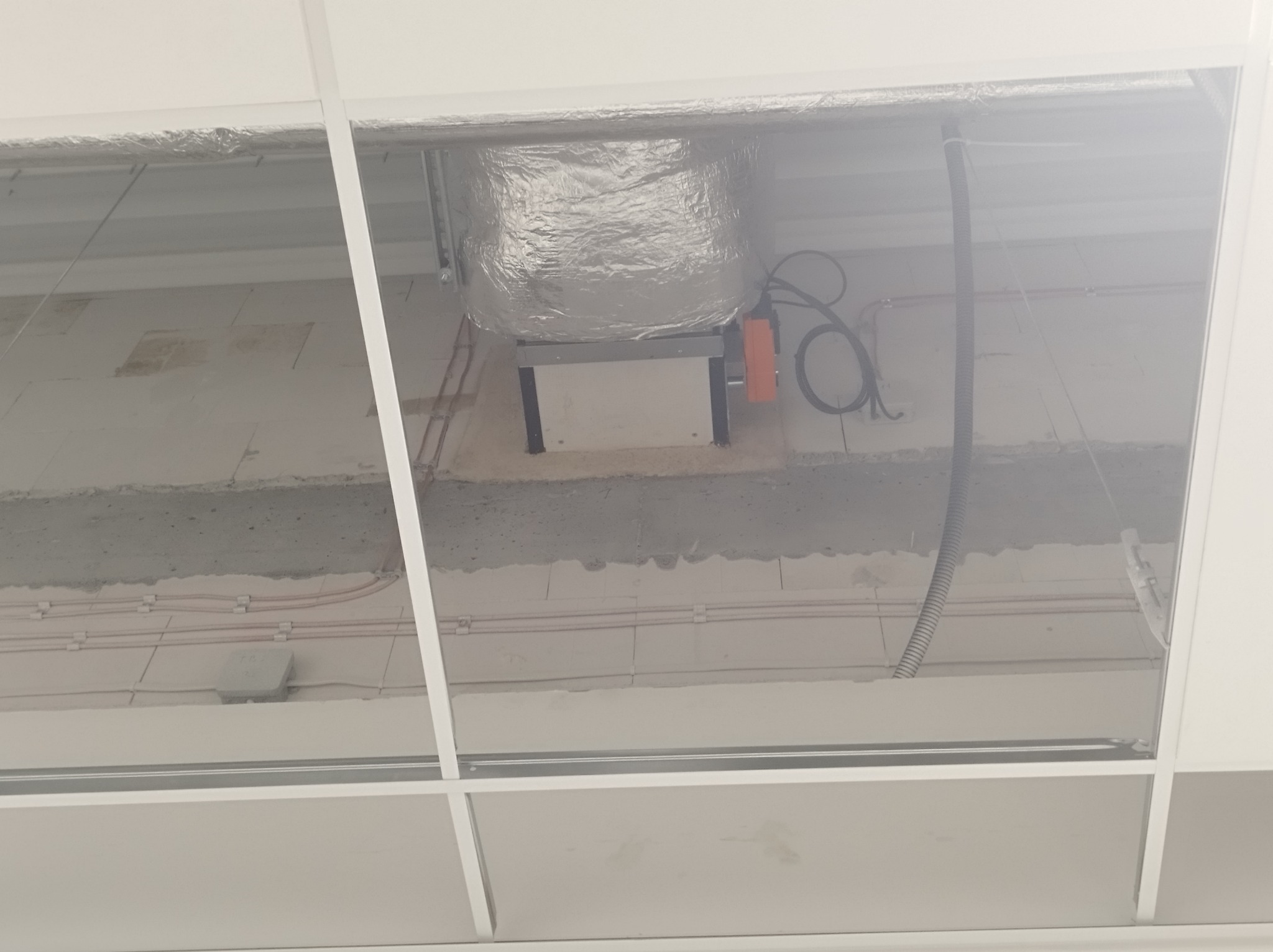 Widok w przestrzeni nad sufitem podwieszanym, widoczny kanał wentylacyjny w srebrnej izolacji, przewody elektryczne i rury instalacyjne.