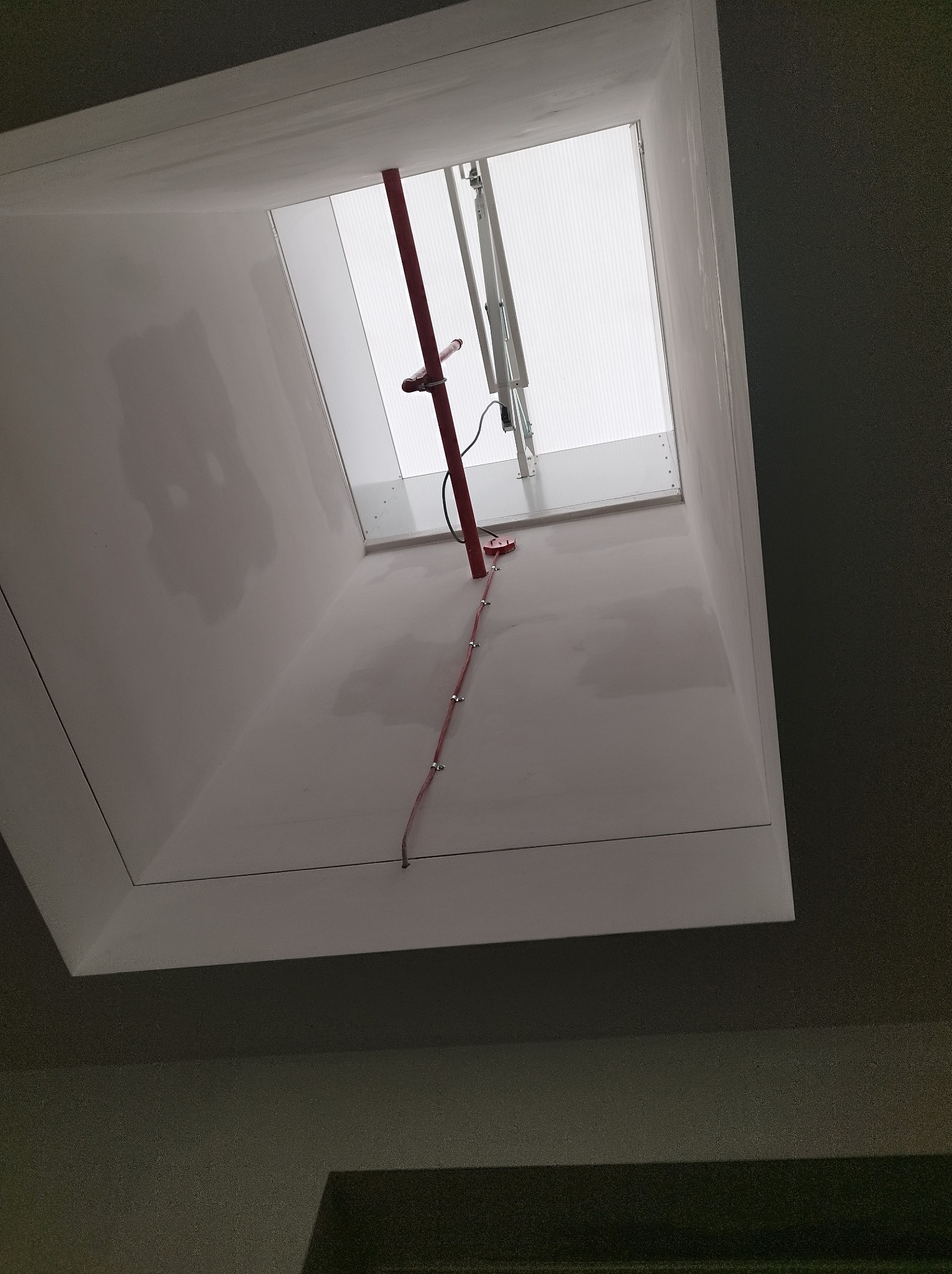 Wnętrze szybu okiennego połaciowego z widoczną konstrukcją okna i poprowadzoną czerwoną rurką instalacyjną z mocowaniami.