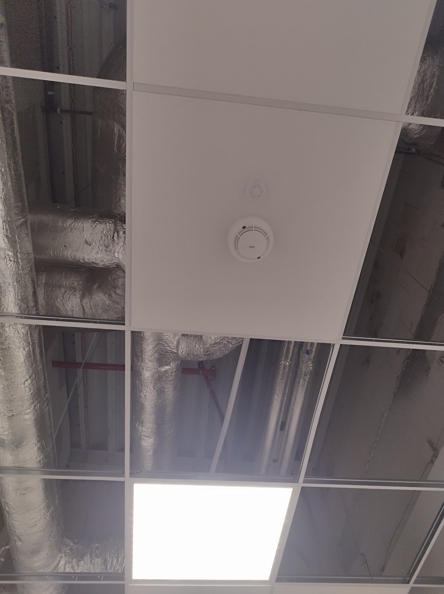 Sufit podwieszany z widocznymi instalacjami wentylacyjnymi w folii aluminiowej, czujnikiem dymu i panelem oświetleniowym LED.