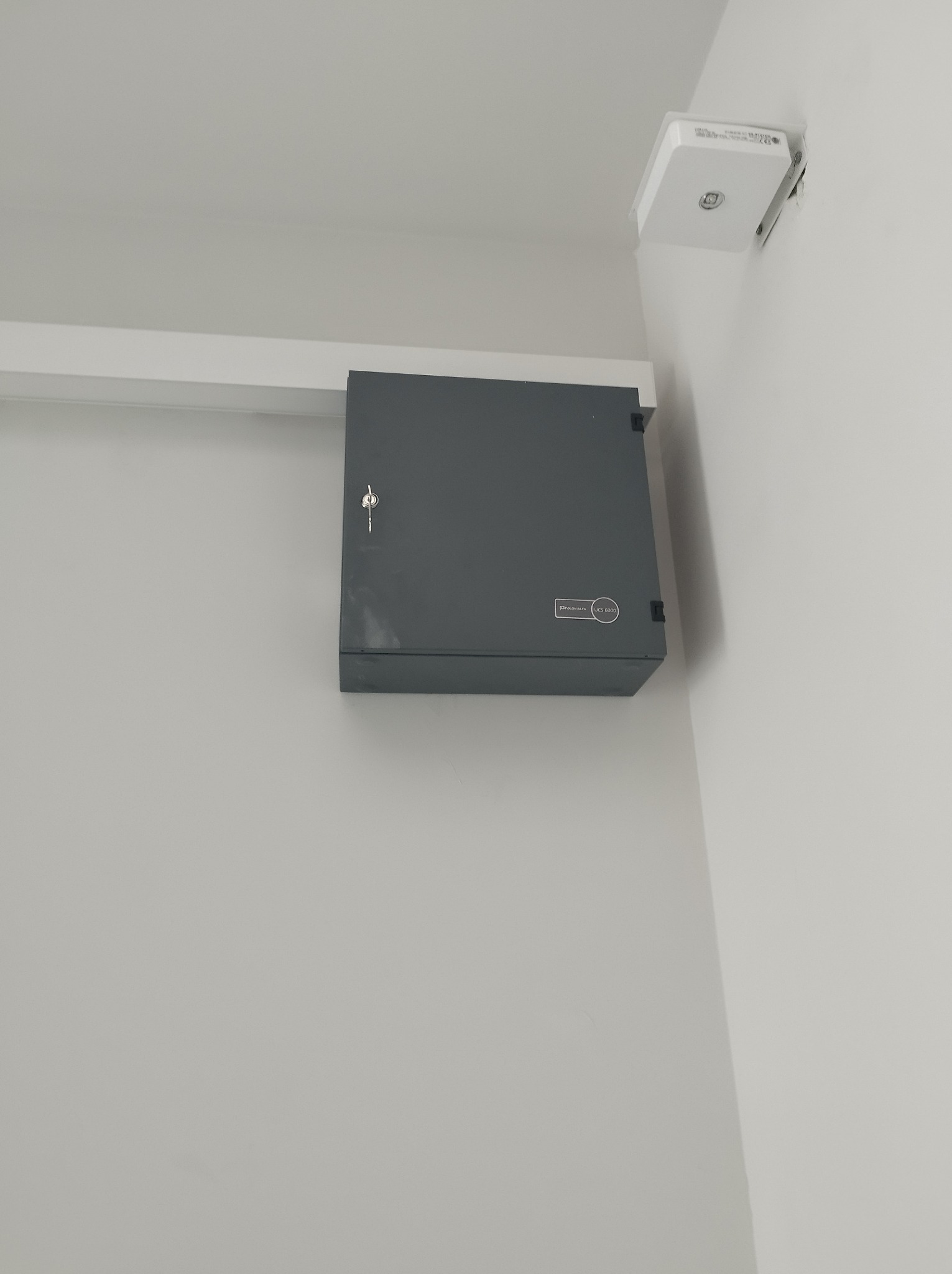 Szara skrzynka elektryczna z kluczem zamontowana pod sufitem, obok biały czujnik na ścianie.