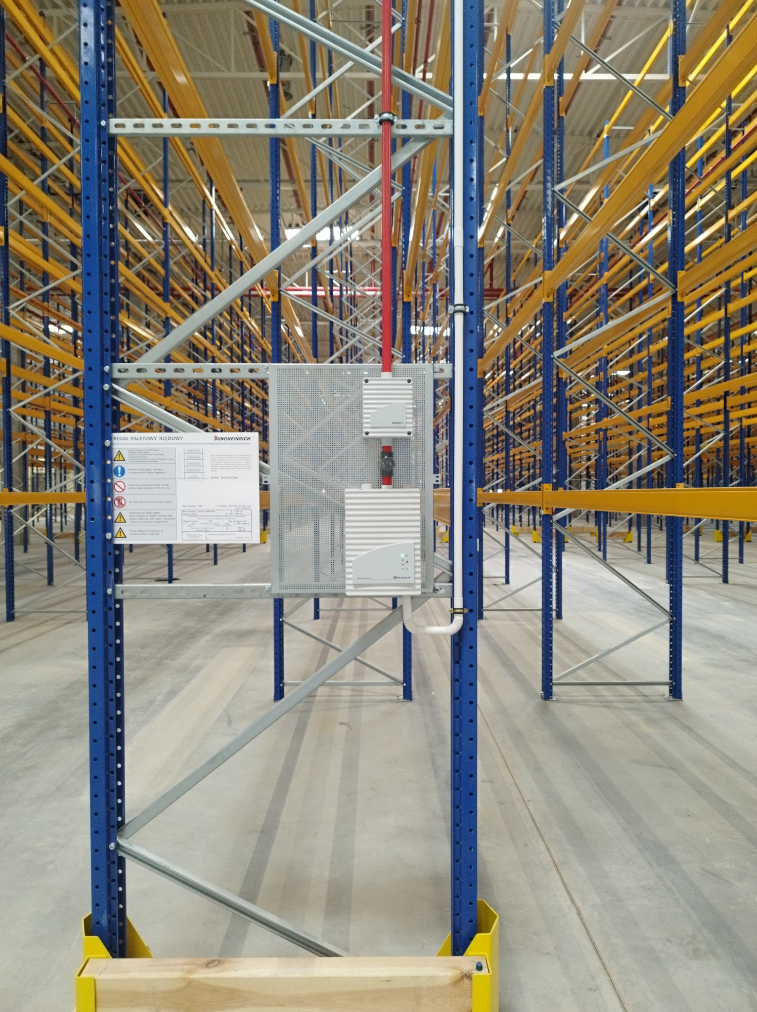 System regałów paletowych z instalacją przeciwpożarową i tabliczką informacyjną, widok z perspektywy posadzki magazynu.