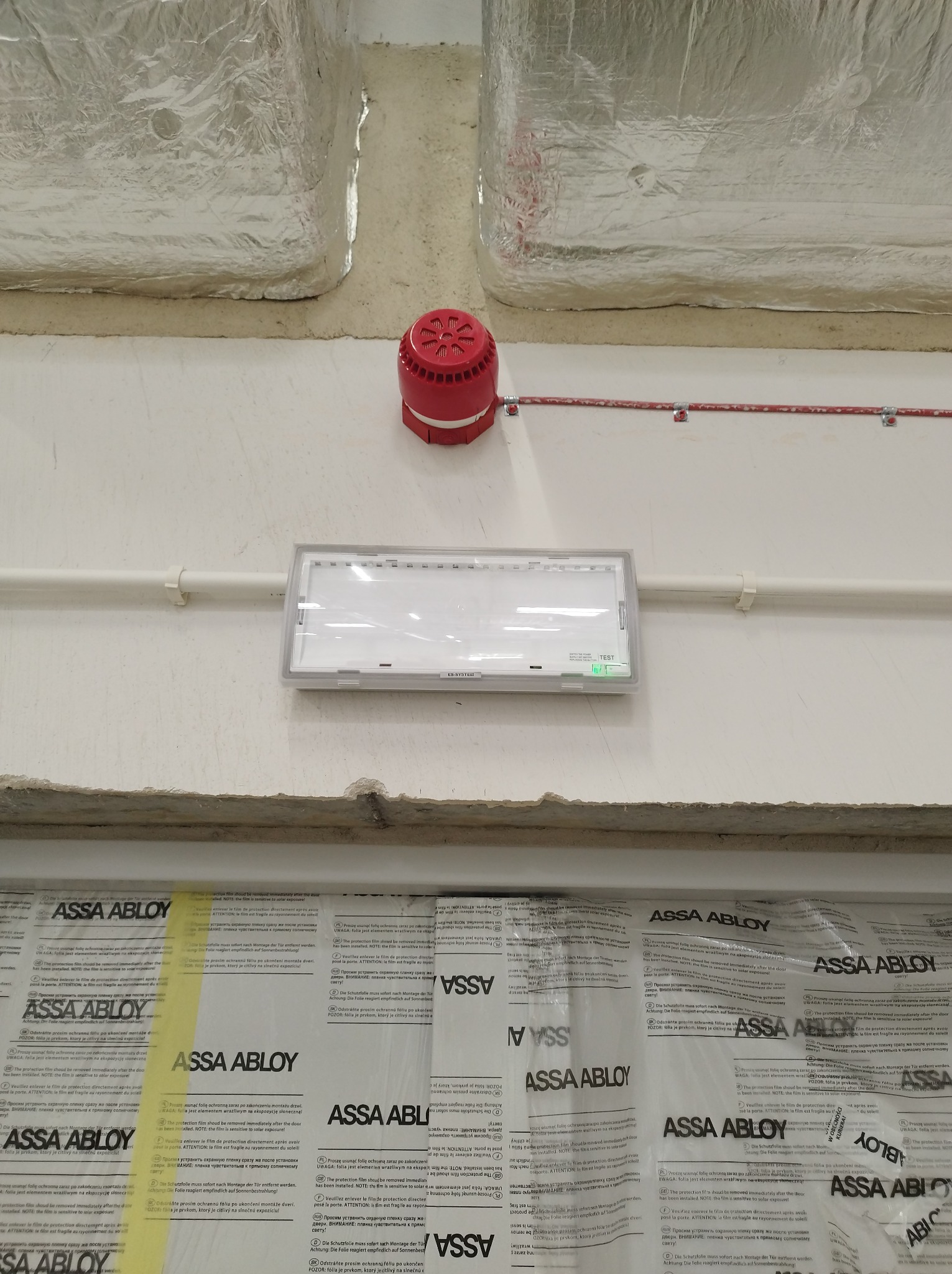 Czerwony sygnalizator alarmowy i biała oprawa oświetleniowa zamontowane na ścianie, widoczne folie ochronne na dole ściany.