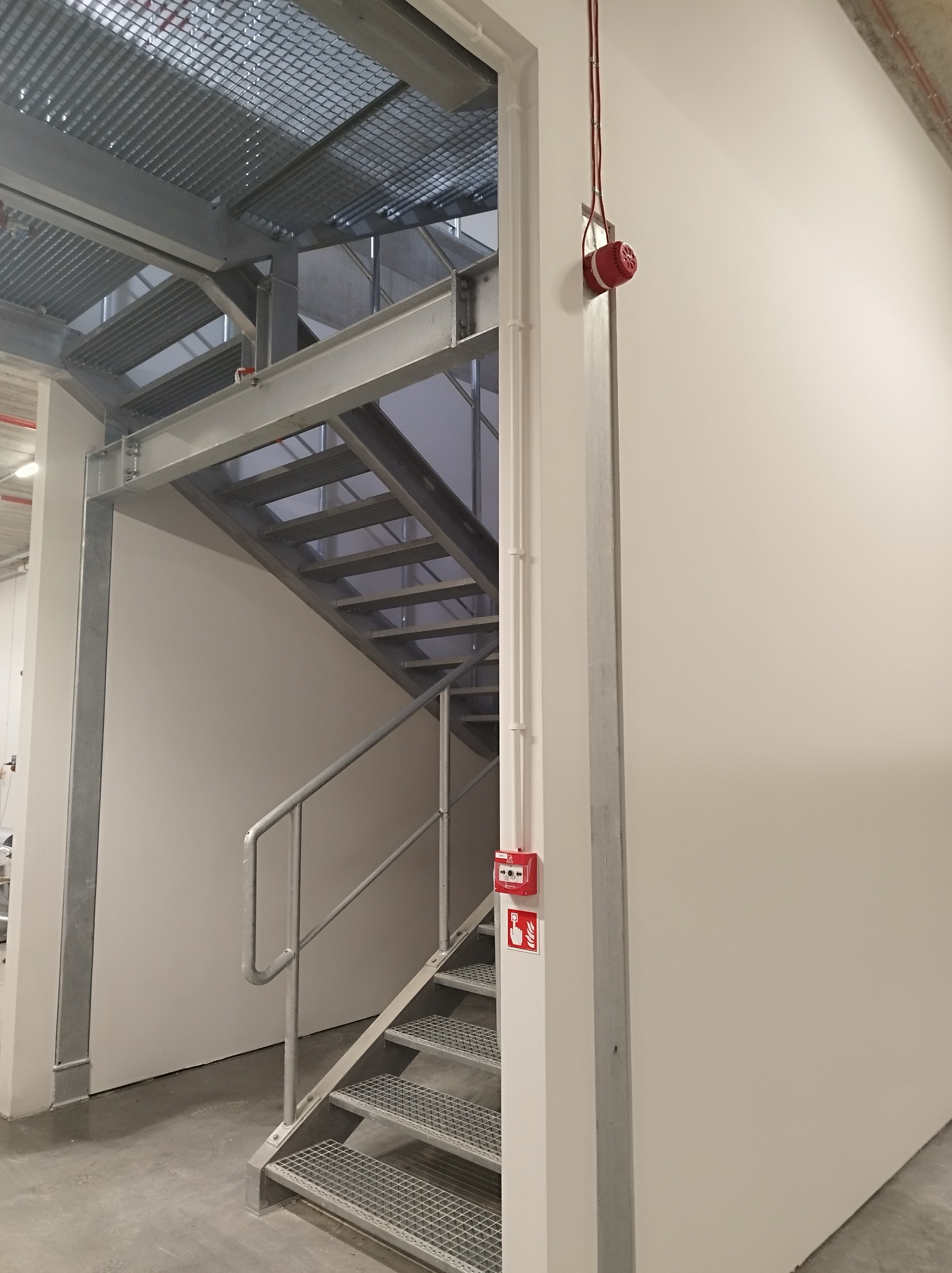 Metalowe schody ewakuacyjne z kratownicowymi stopniami i barierką, widoczny sygnalizator pożarowy na ścianie i przewody elektryczne w listwach.