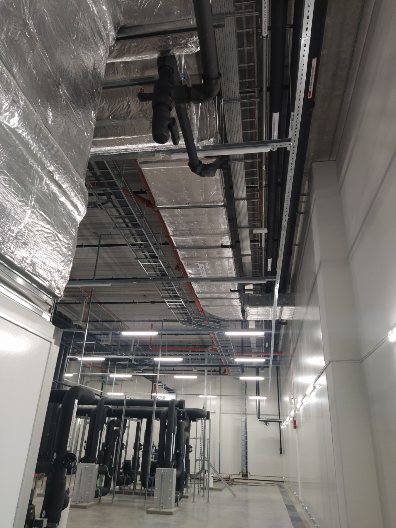 Wnętrze hali przemysłowej z widocznymi instalacjami: rury, kable w korytach, oświetlenie LED i izolacja termiczna na suficie.