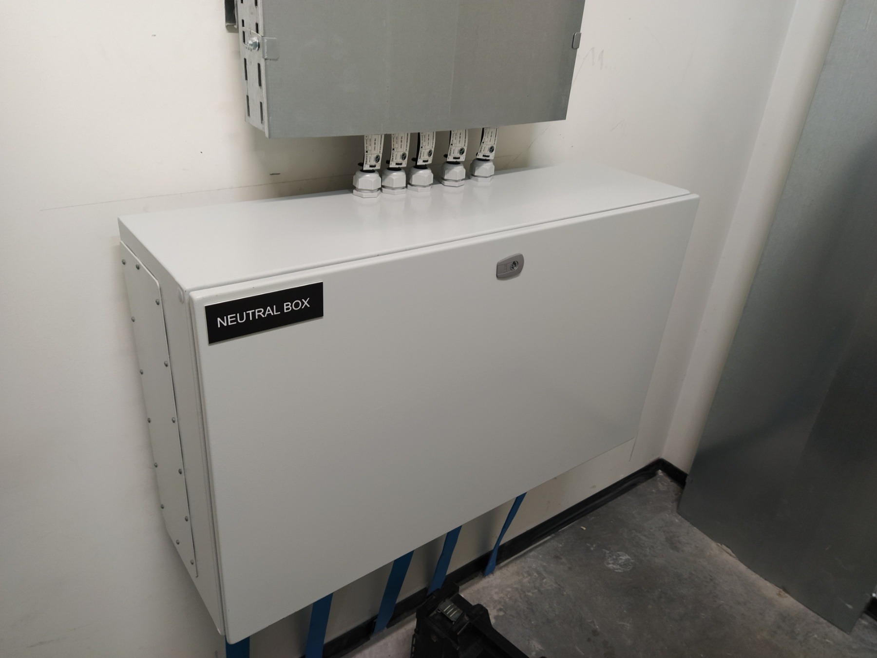 Biała skrzynka neutralna z widocznymi przewodami i złączkami na tle ściany, z etykietą 'NEUTRAL BOX' po lewej stronie.