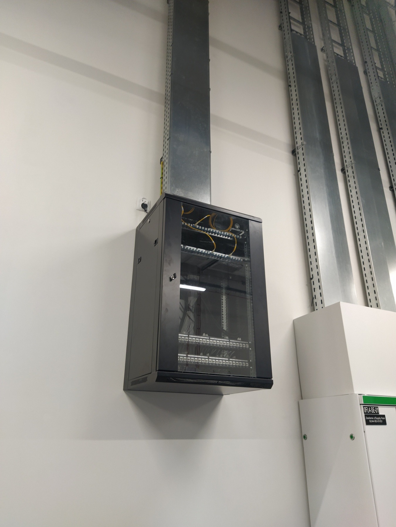 Szara szafa rackowa zamontowana na białej ścianie, z widocznymi kablami i przewodami, z metalową rurą wentylacyjną prowadzącą do góry i perforowanymi szynami montażowymi po prawej stronie.