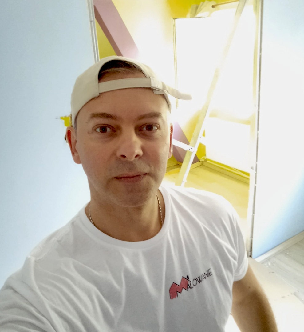 Mężczyzna w czapce i koszulce z napisem 'Malowanie' na tle pomalowanego pokoju z drabiną