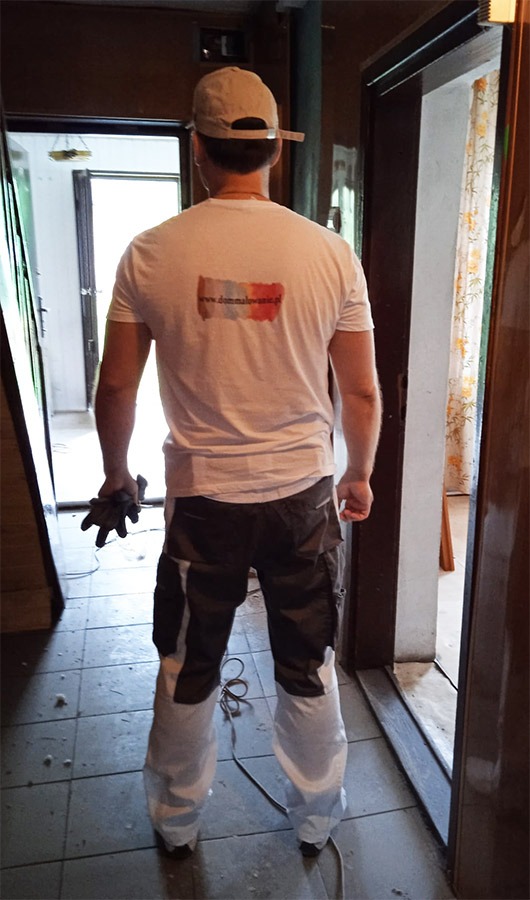 Mężczyzna w roboczym stroju z logo firmy malarskiej na koszulce, stojący w korytarzu w trakcie remontu, trzymający rękawice robocze.