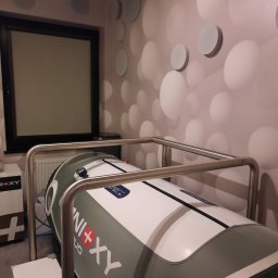 CENTRUM PSYCHOSOMATYKI - Komora hiperbaryczna w pomieszczeniu z designerską ścianą w kropki, widoczny metalowy stelaż i okno z żaluzjami w tle.