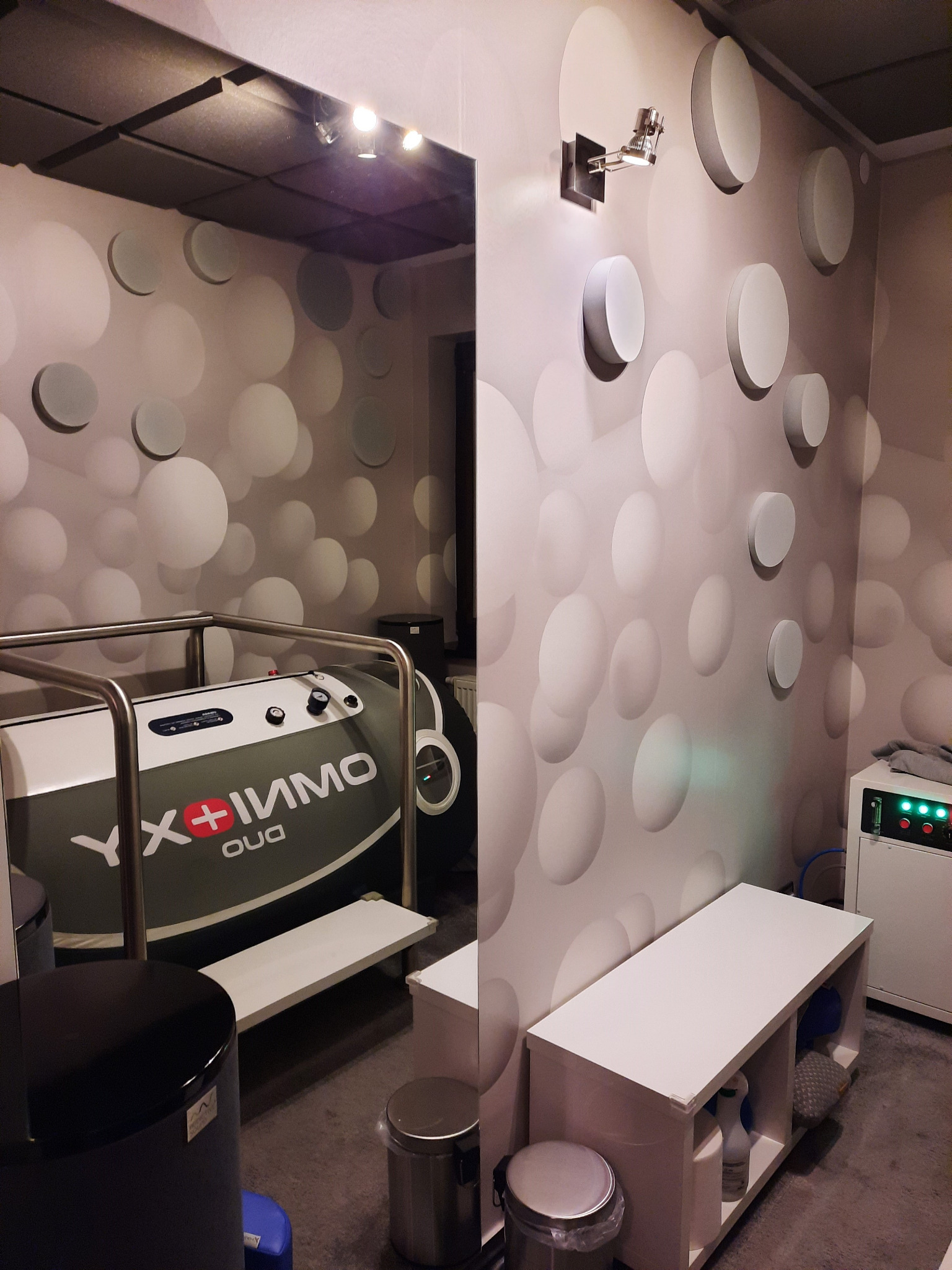 Wnętrze gabinetu z kapsułą do hydroterapii z napisem YX+IMMO OUA, widok z perspektywy lustra, ściana z wypukłymi, okrągłymi dekoracjami, biała szafka i urządzenie z zielonymi diodami.