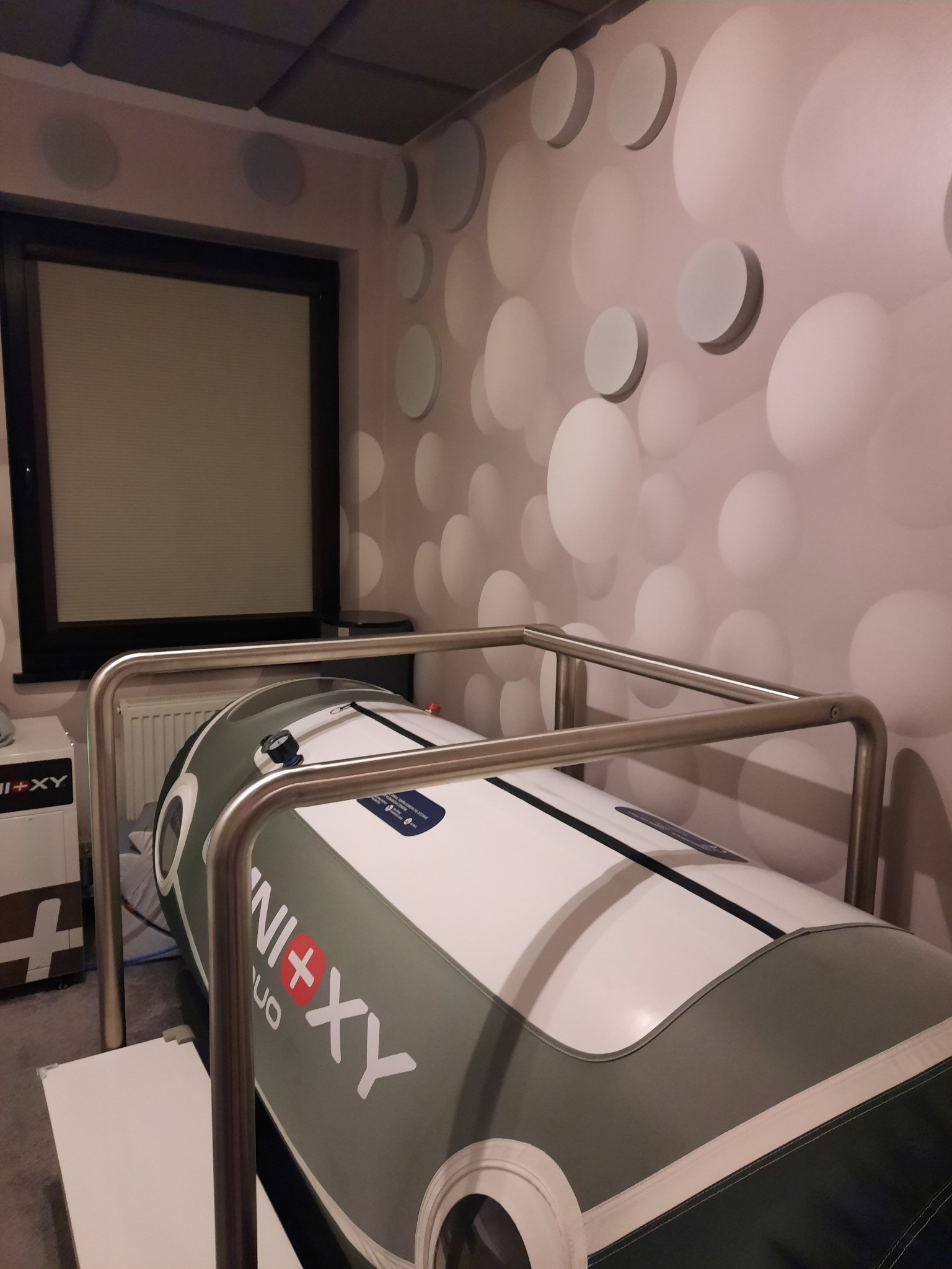 Komora hiperbaryczna w pomieszczeniu z designerską ścianą w kropki, widoczny metalowy stelaż i okno z żaluzjami w tle.